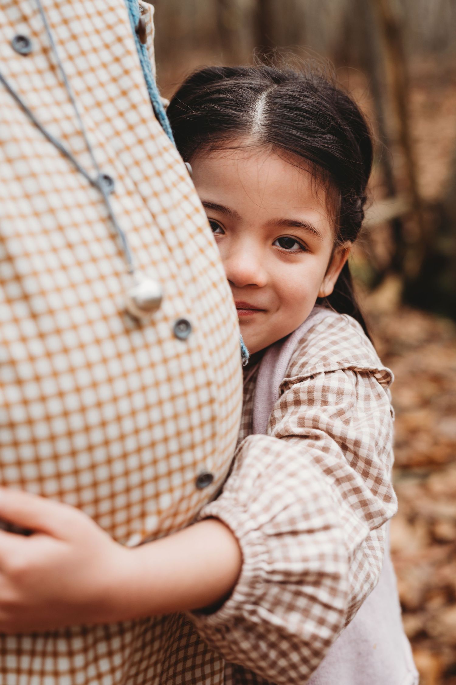 A young girl hugs her mother's pregnant belly while smiling at the camera in an autumn forest.