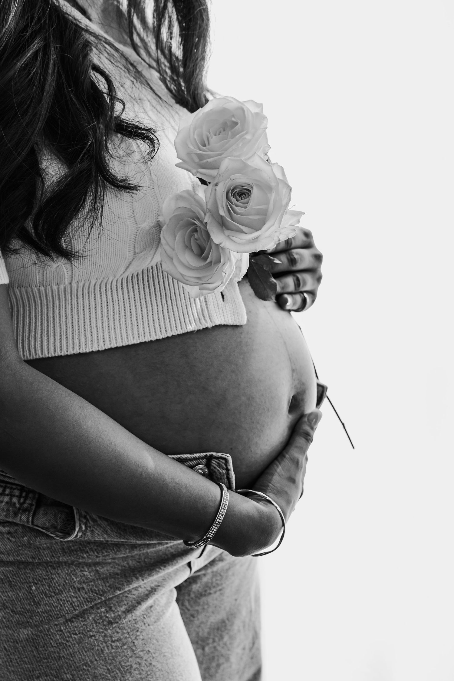 Black and white maternity photo of pregnant woman holding white roses against belly.
