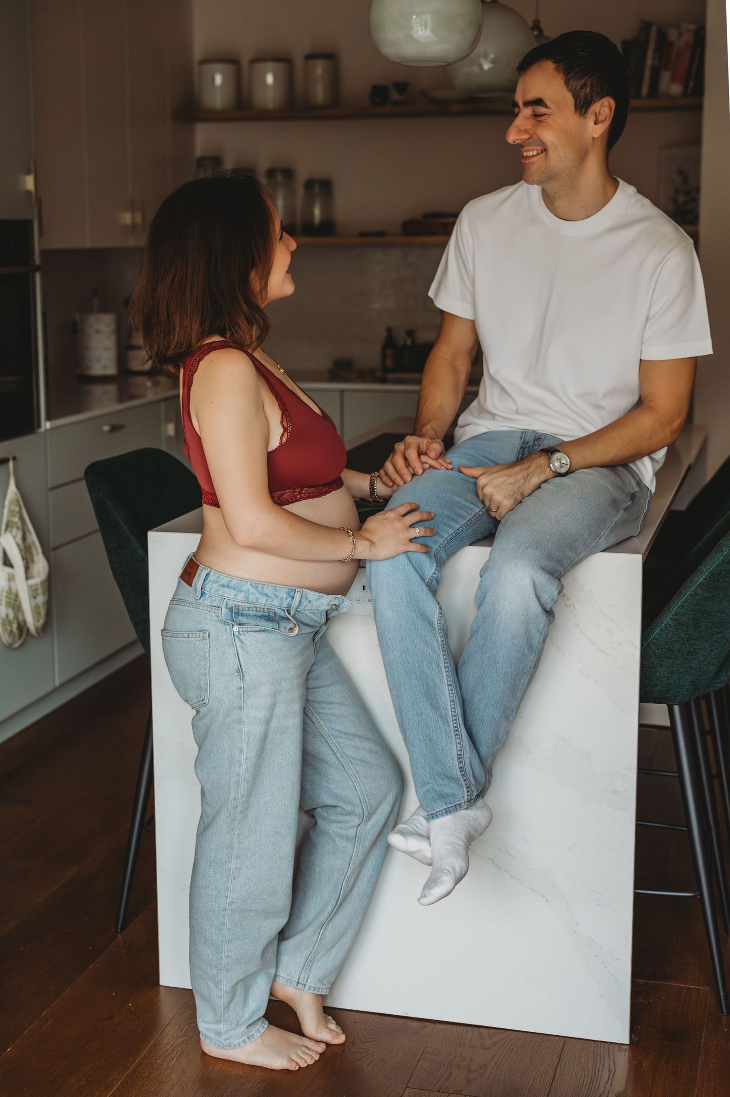 Pregnant woman in red crop top and jeans smiling at partner seated on white kitchen island.