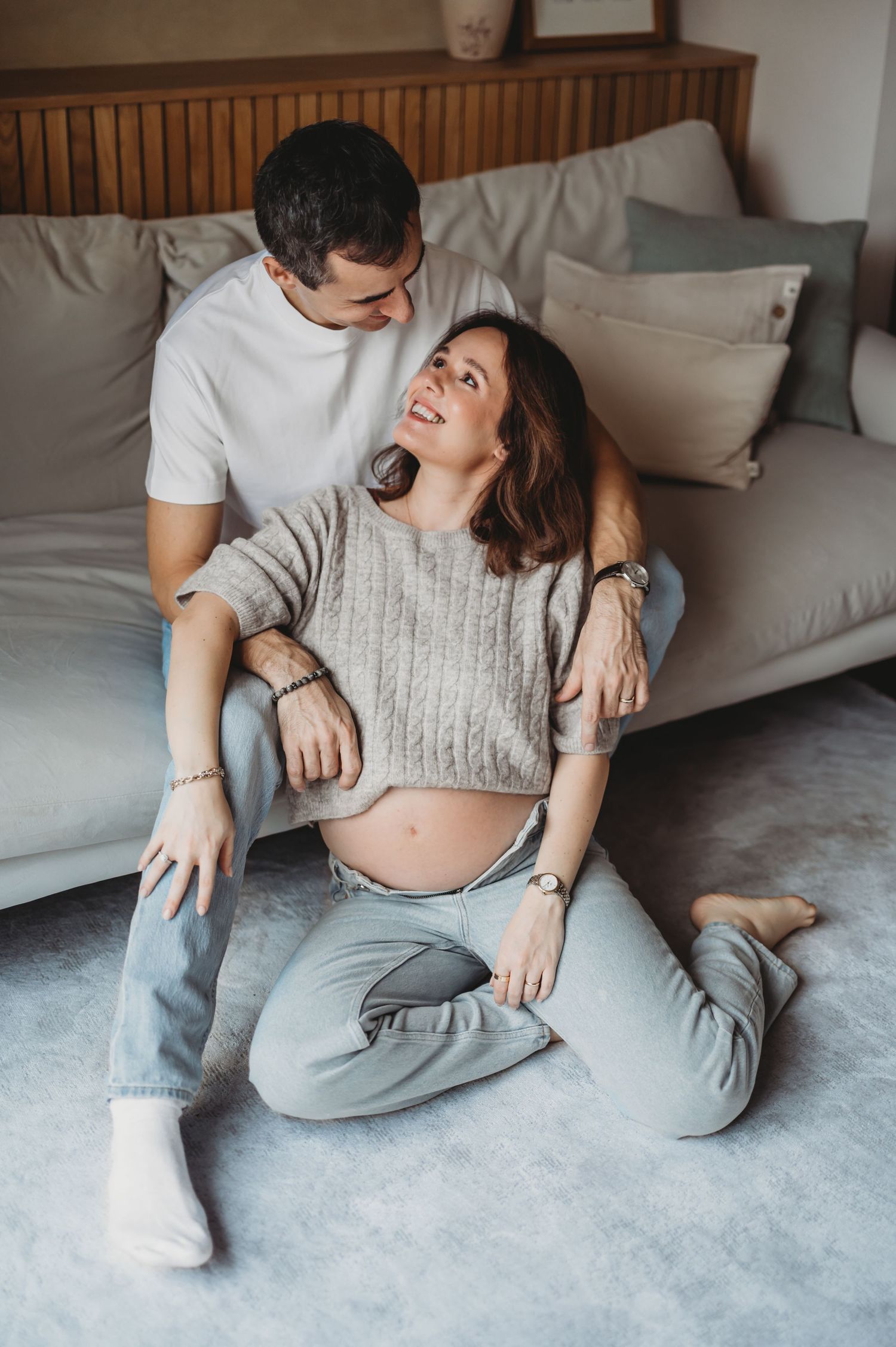 A happy pregnant woman sits on the floor leaning against a sofa while her partner embraces her warmly.