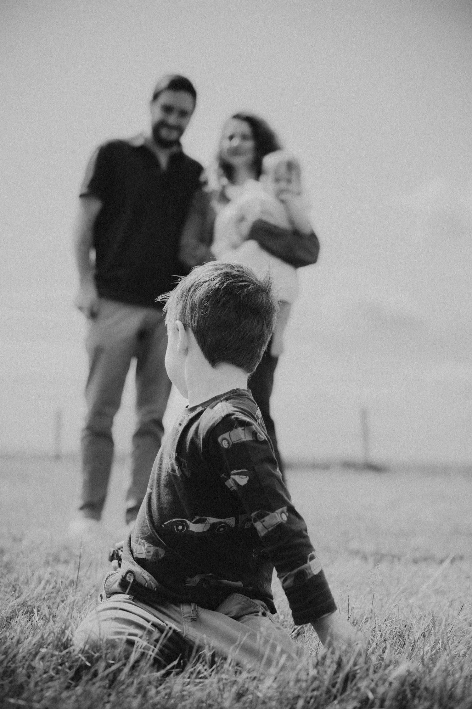 Black and white family portrait with young boy in foreground and parents holding baby in a field.