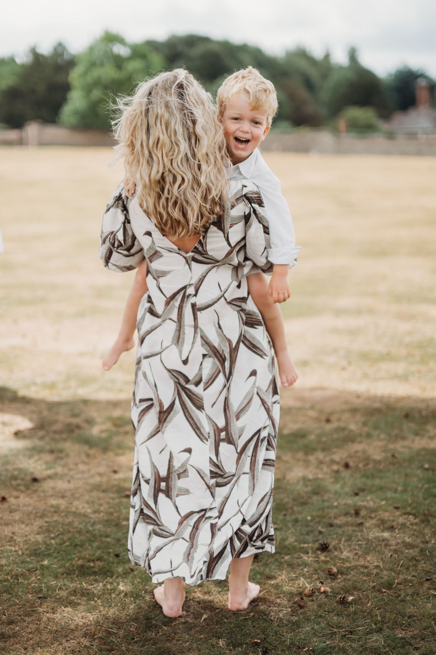 A curly-haired woman in a leaf-print dress carries a laughing blonde toddler across a grassy field.