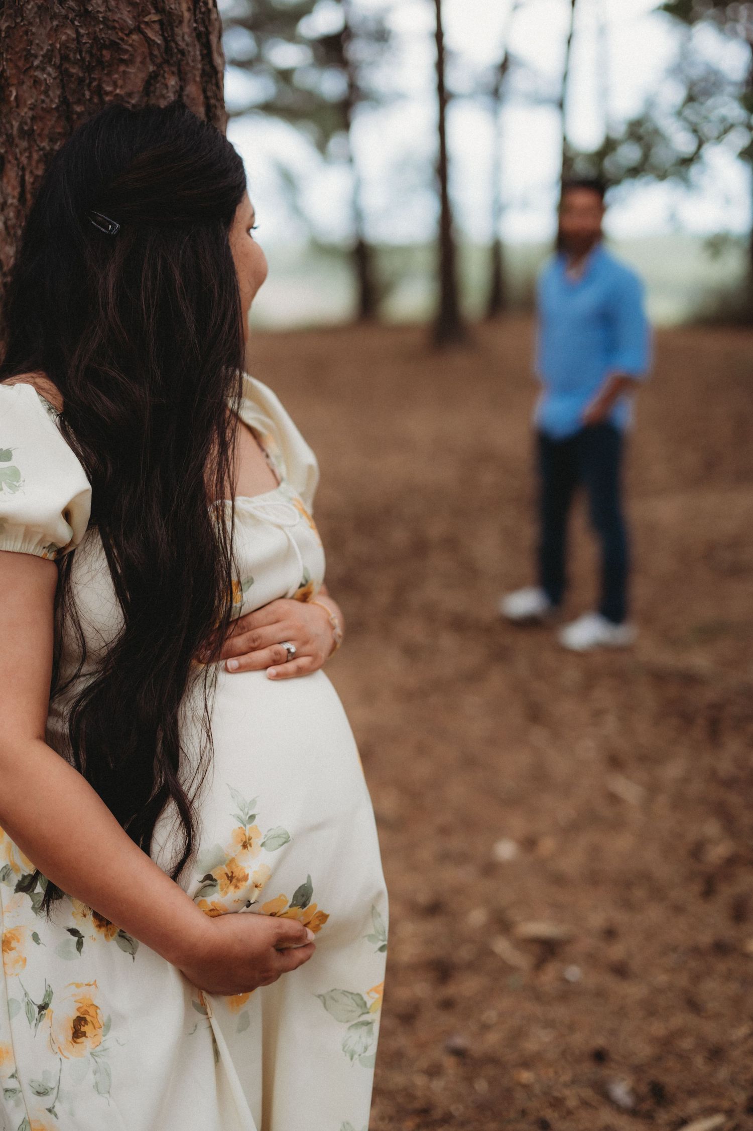 Pregnant woman in floral dress with partner blurred in forest background.
