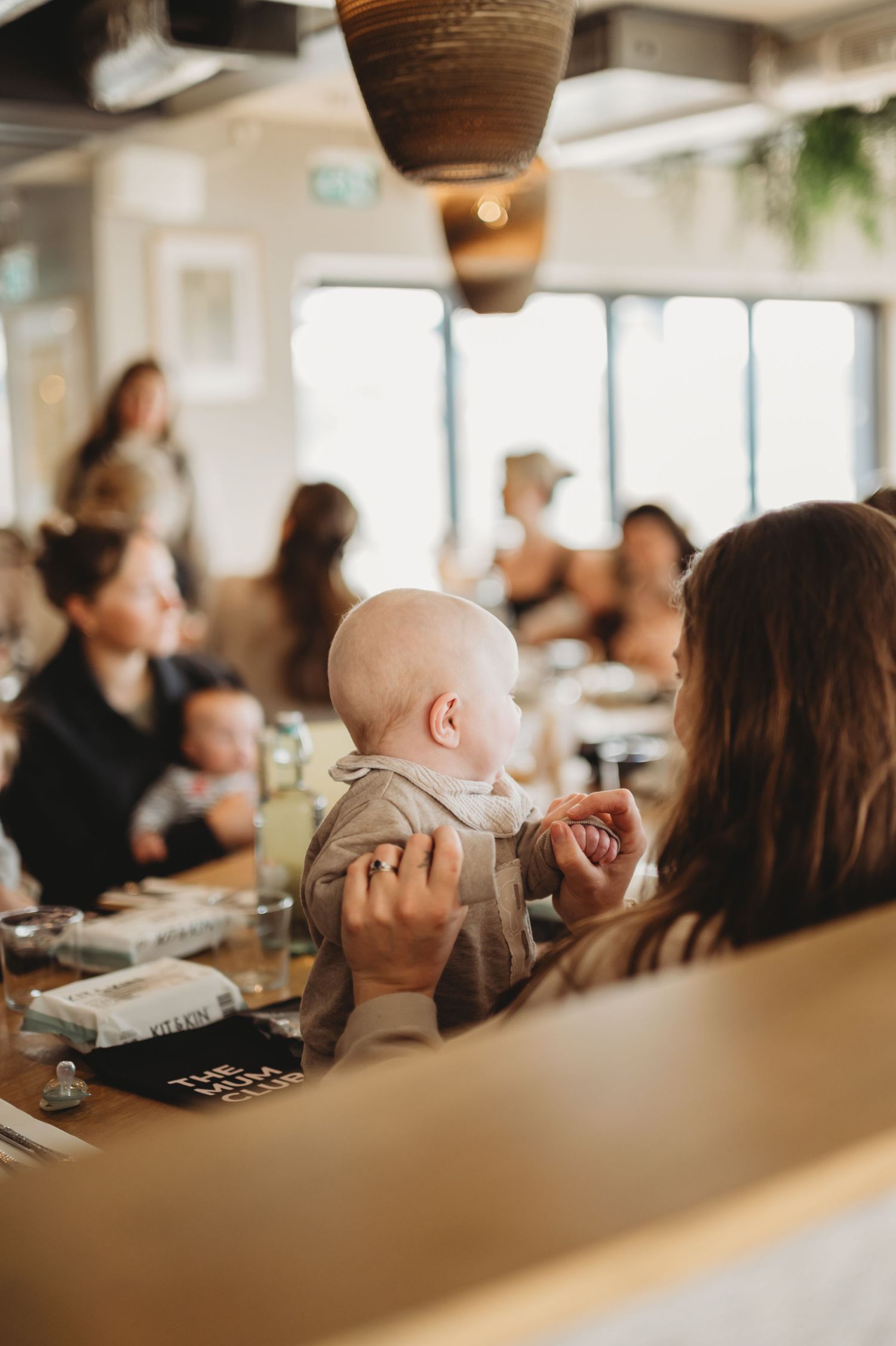 Mother holding a young baby at a busy indoor cafe gathering with other parents.