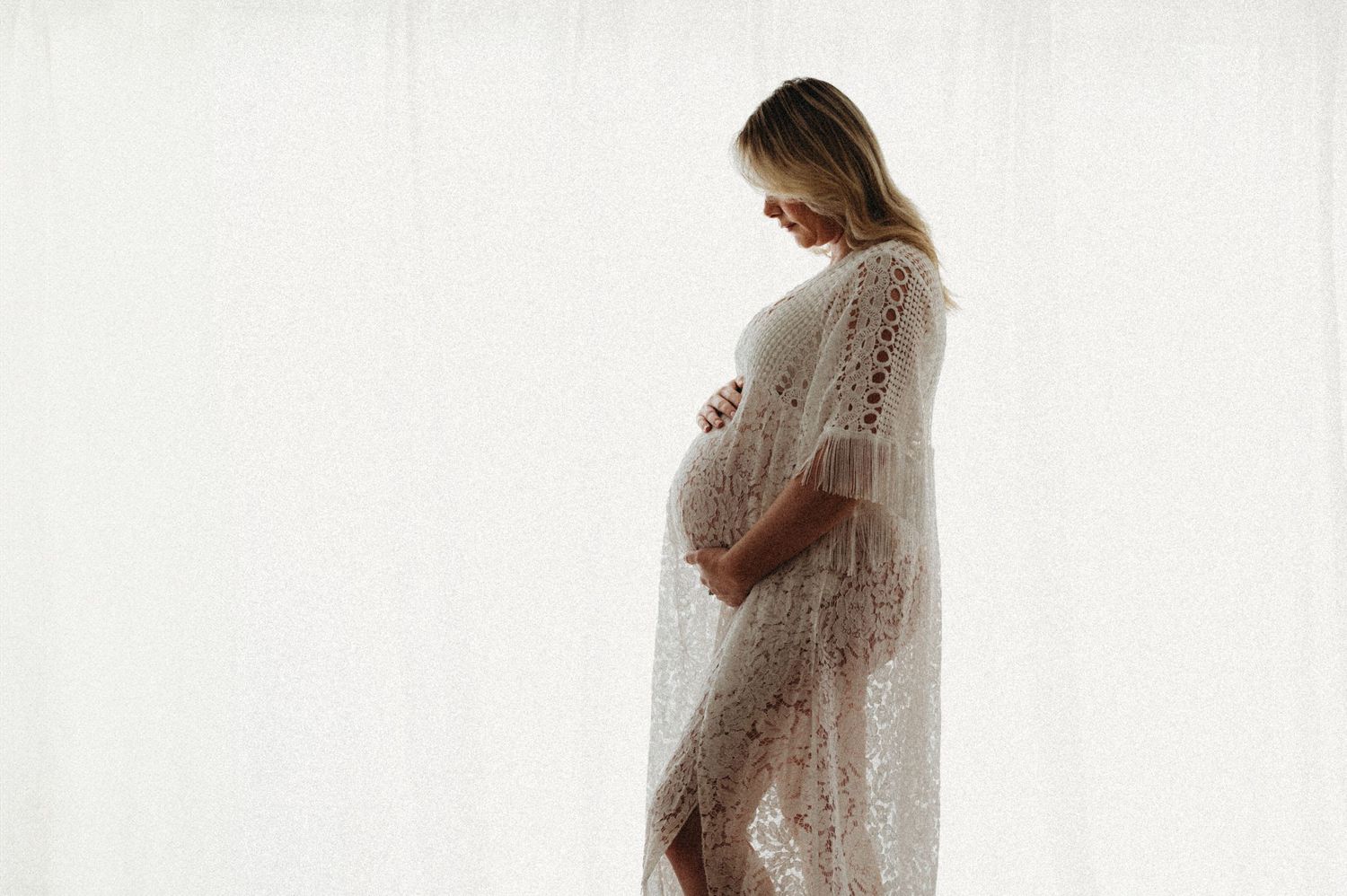 Pregnant woman in a delicate lace robe gently holds her baby bump against a white backdrop.
