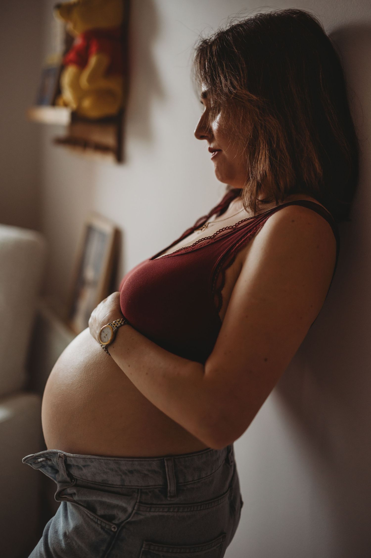 Pregnant woman in red crop top and jeans cradling her baby bump against a wall in a moody indoor setting.