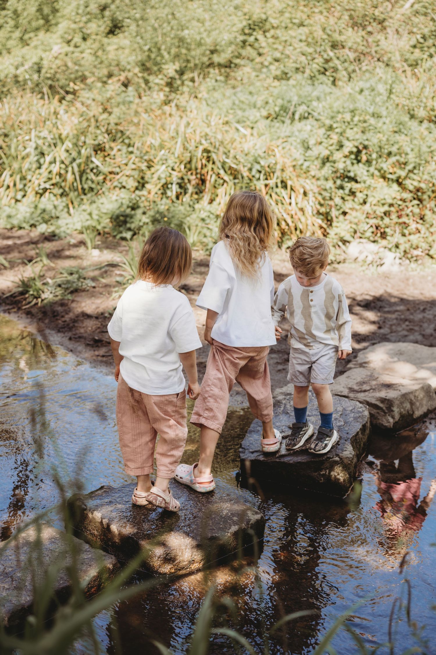 Three children in neutral clothing stepping on rocks across a shallow stream in nature.
