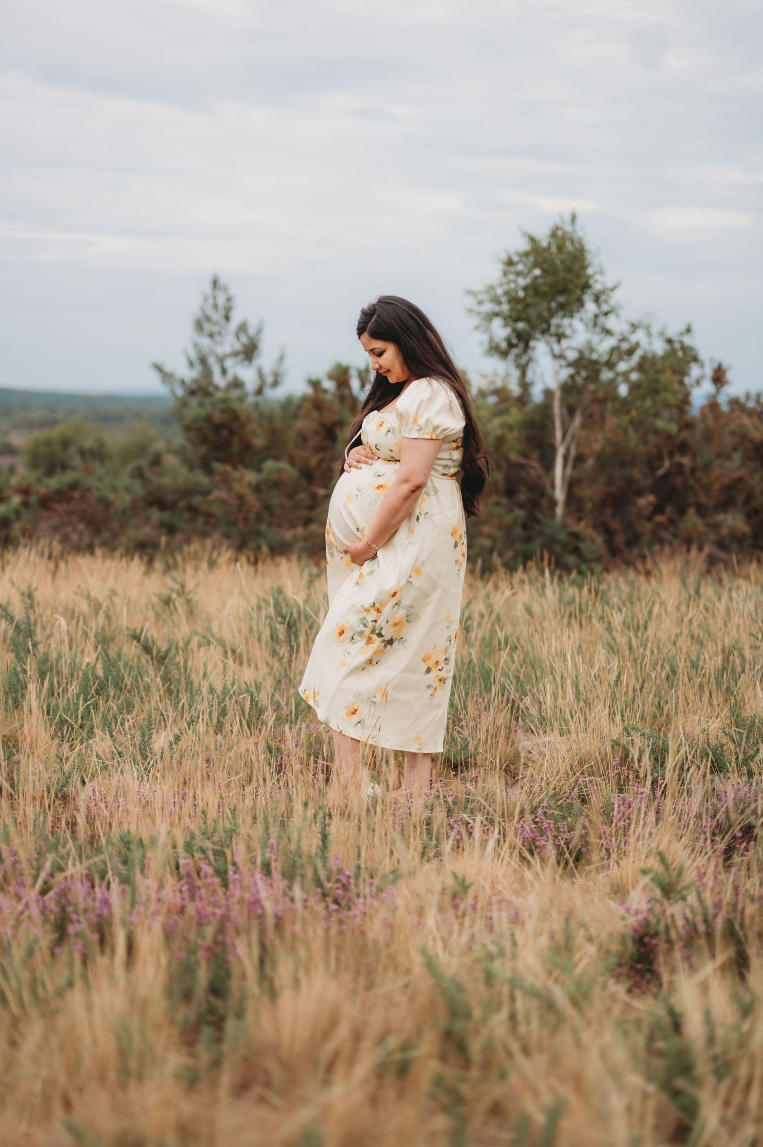 Pregnant woman in floral dress standing in golden meadow field outdoors.