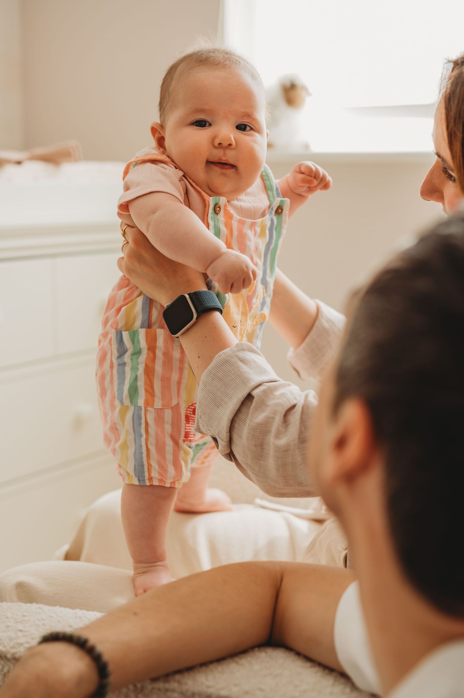 Happy baby in colorful striped romper standing with parent support in bright nursery room.