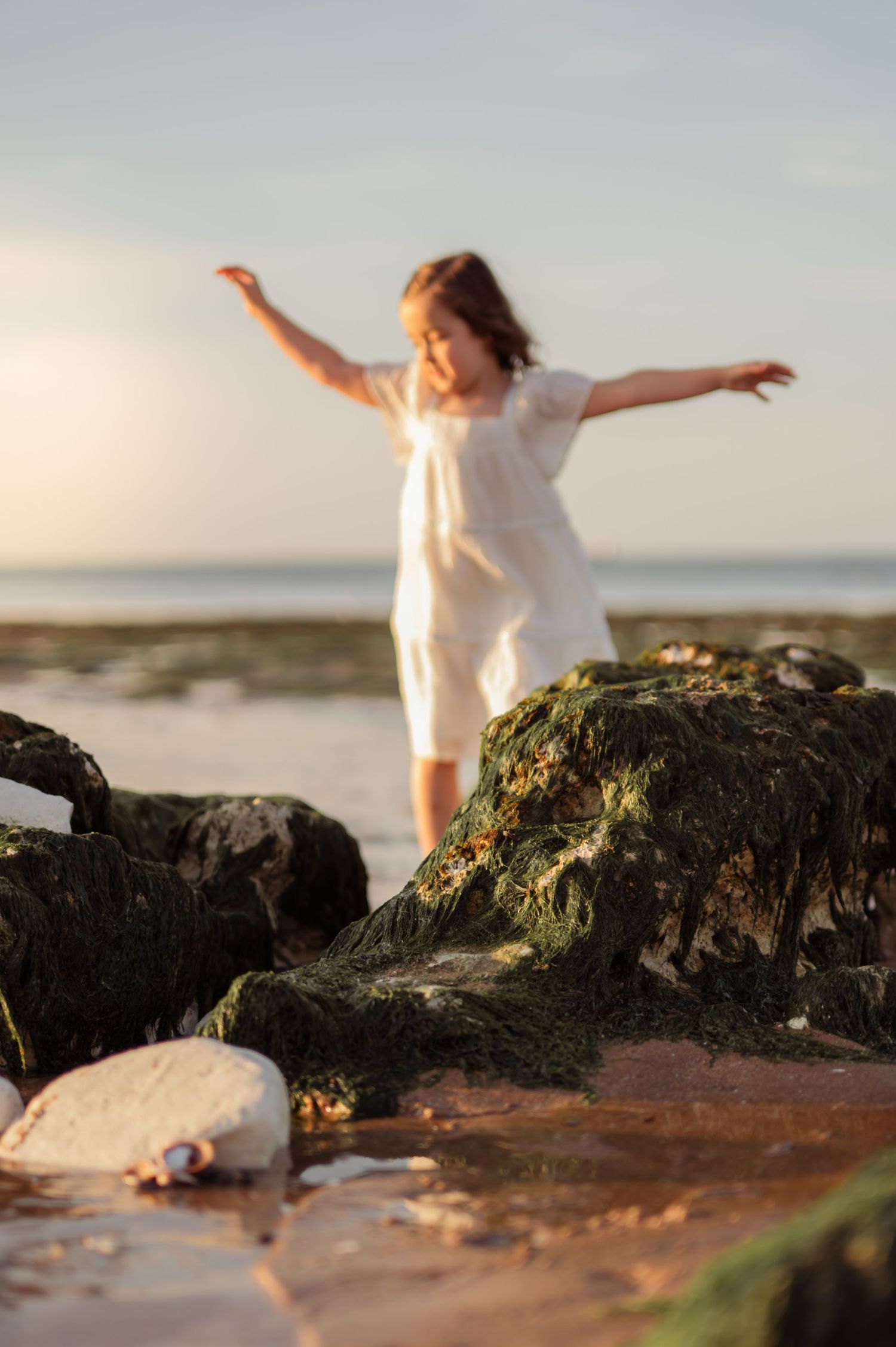 Young girl in white dress balancing on mossy rocks at the beach during golden hour sunset.
