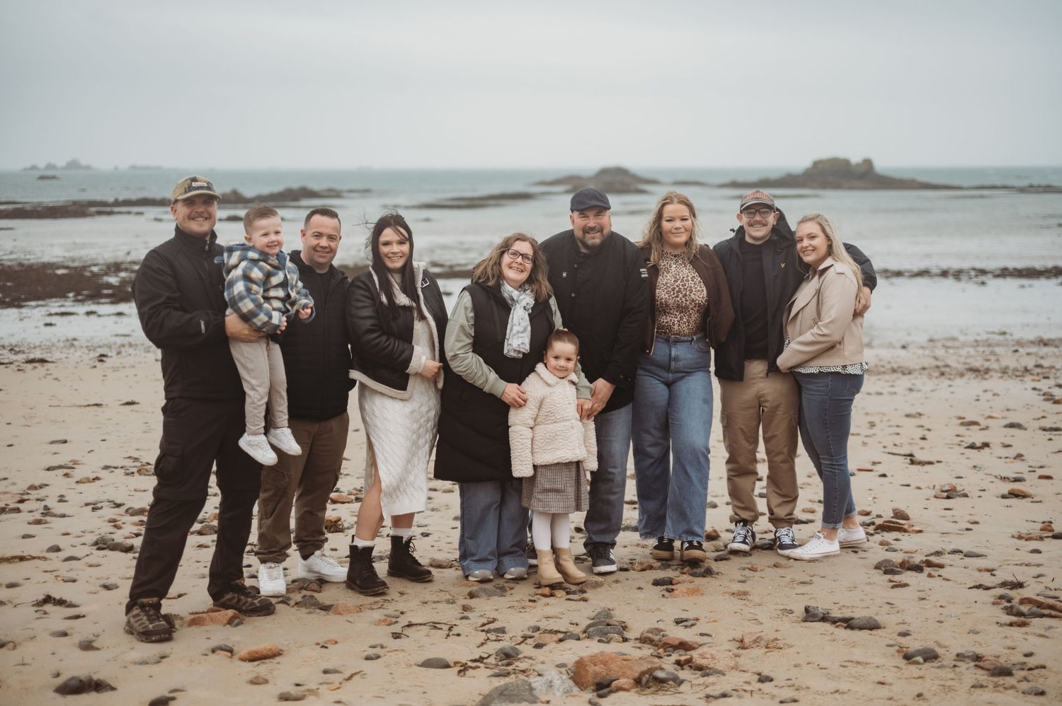 Extended family group portrait of nine people posing together on a rocky beach on an overcast day.