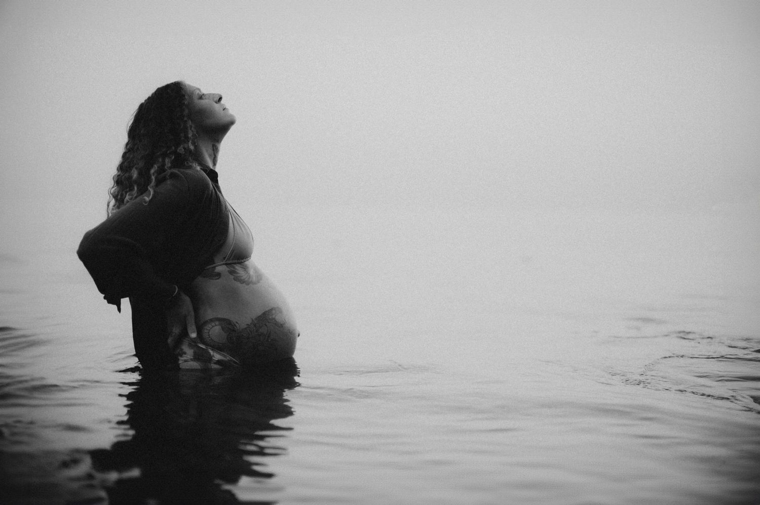 Pregnant woman standing in misty water, head tilted back, cradling baby bump in black and white maternity photo.