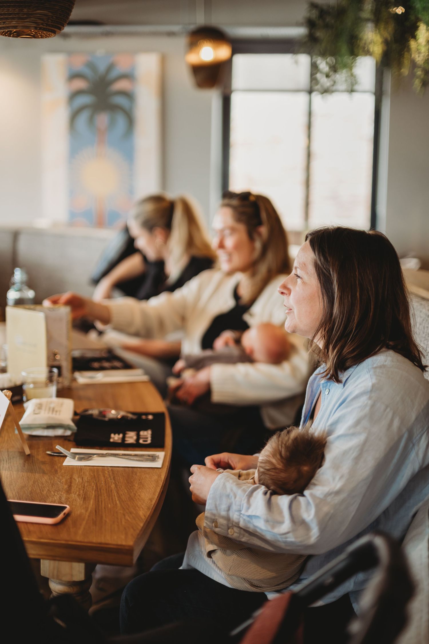 Women sitting at a restaurant table holding babies during a Mum Club gathering event.