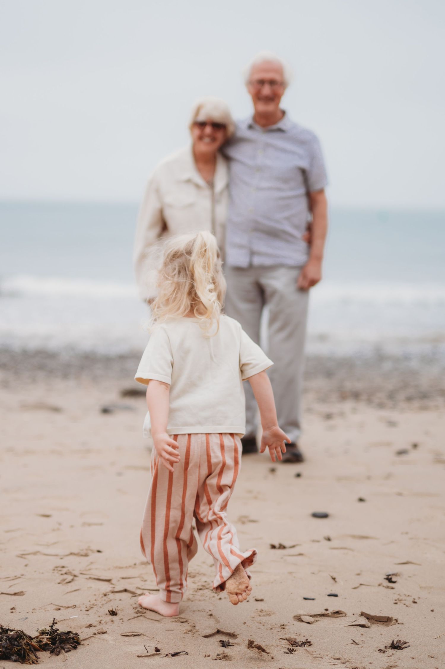 A young child walks toward grandparents standing on a sandy beach by the ocean.