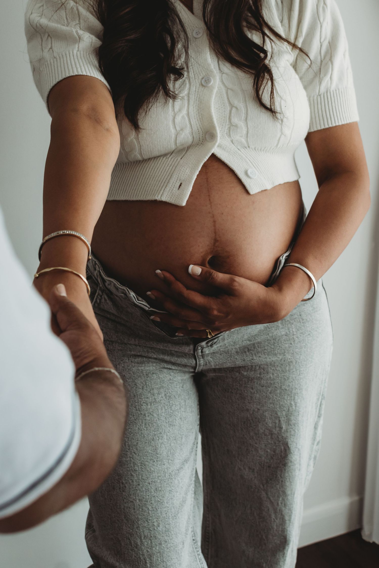 Pregnant woman in white crop top and jeans cradling baby bump indoors.