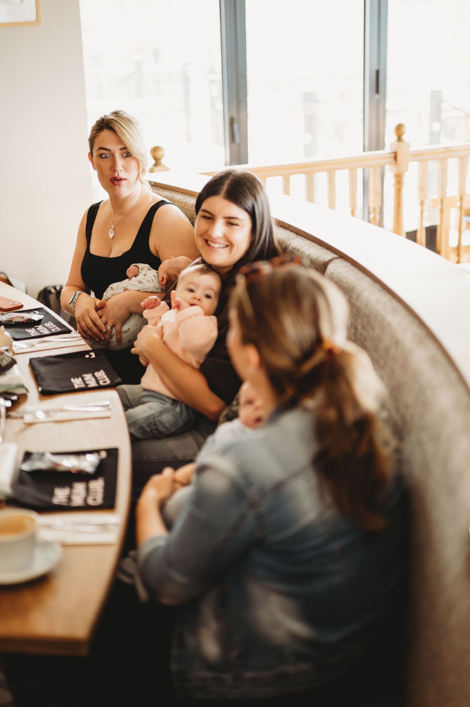 Three mothers with babies socializing in a cozy booth at a bright cafe gathering.