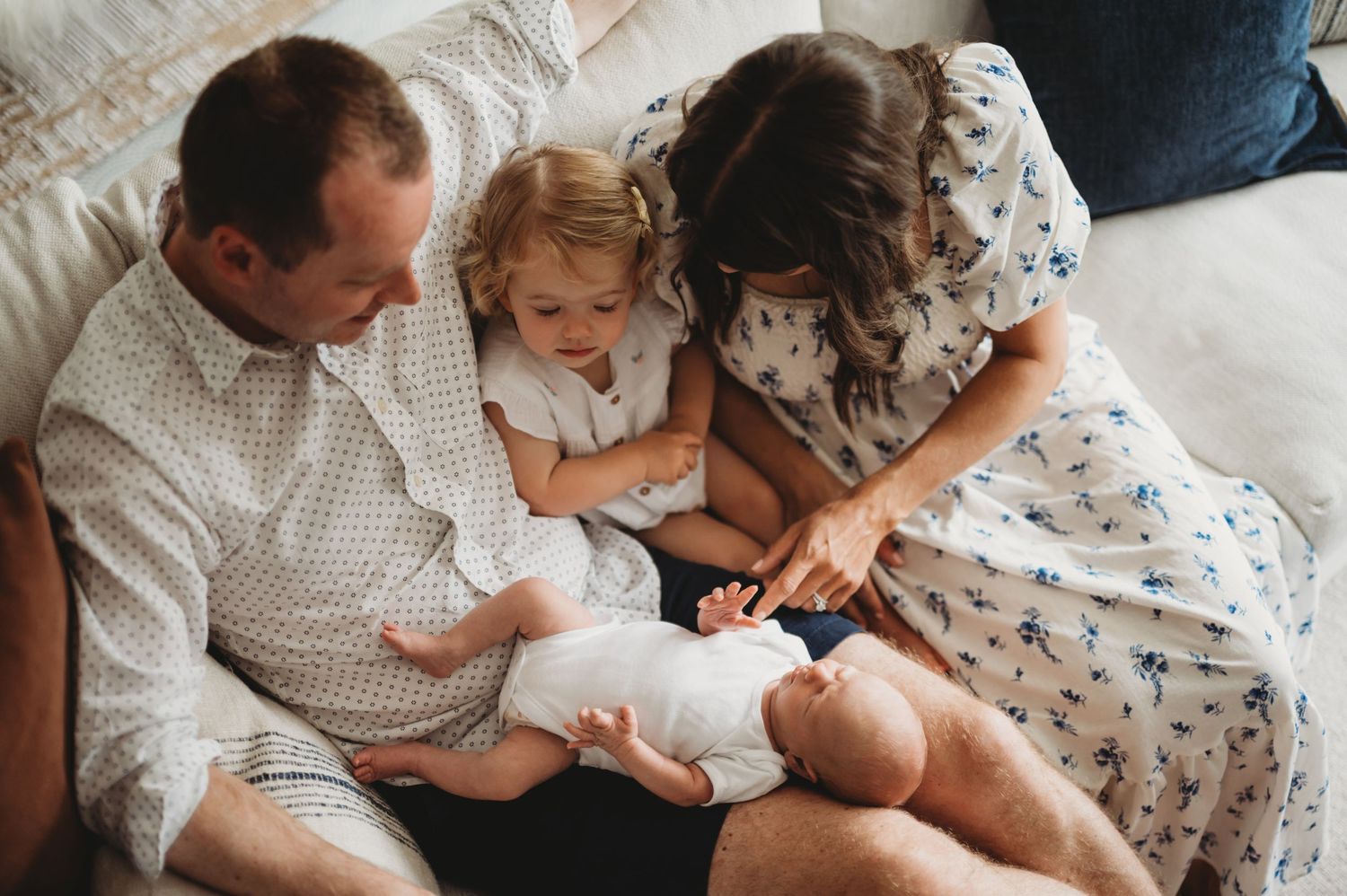 Family of four with parents and toddler admiring newborn baby together on a cozy couch at home.