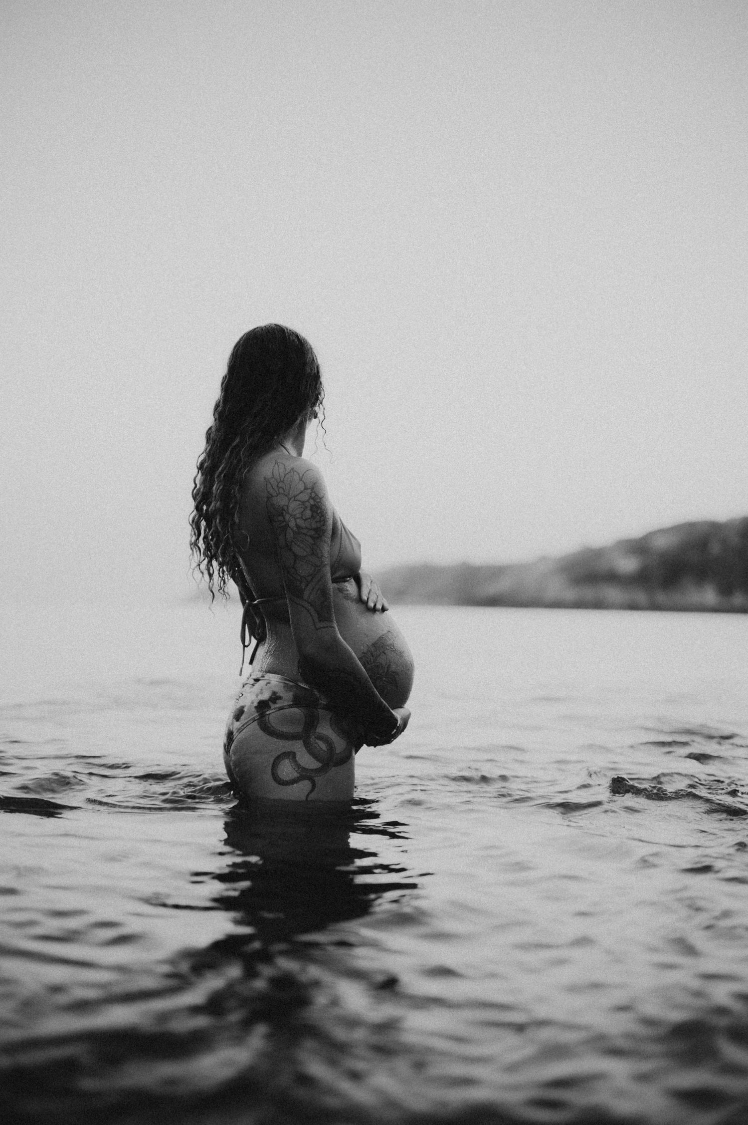 Pregnant woman with tattoos standing in water, black and white maternity photography outdoors.