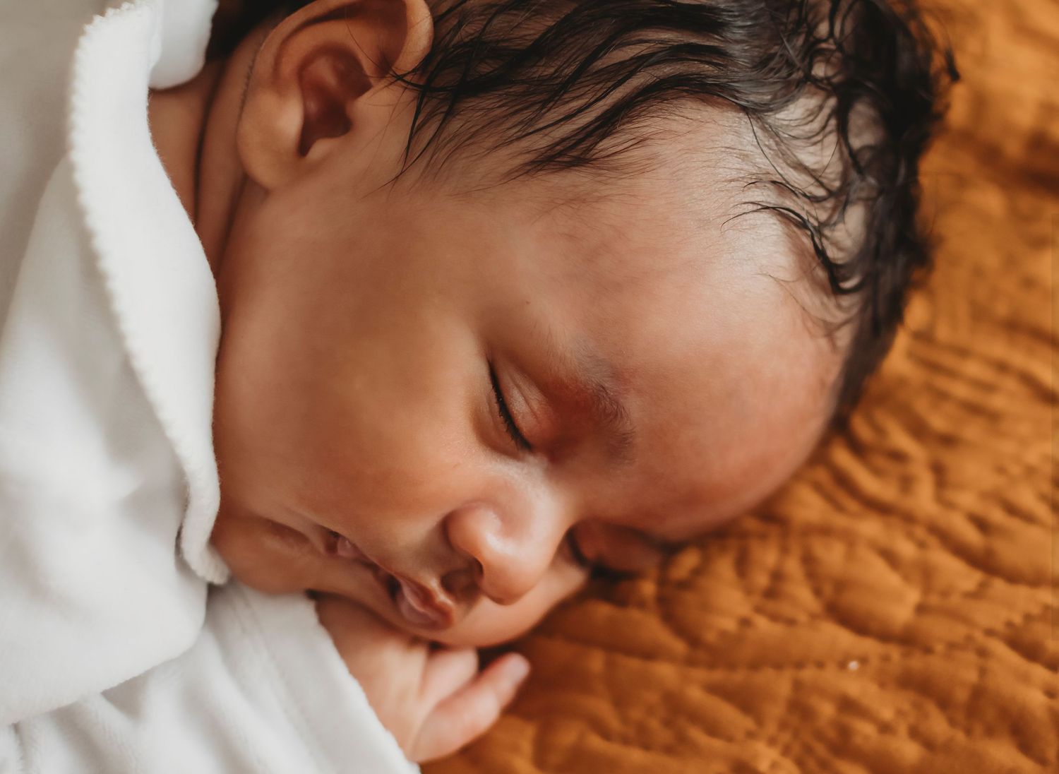 Sleeping newborn baby with dark hair resting peacefully on an orange quilted blanket wrapped in white.