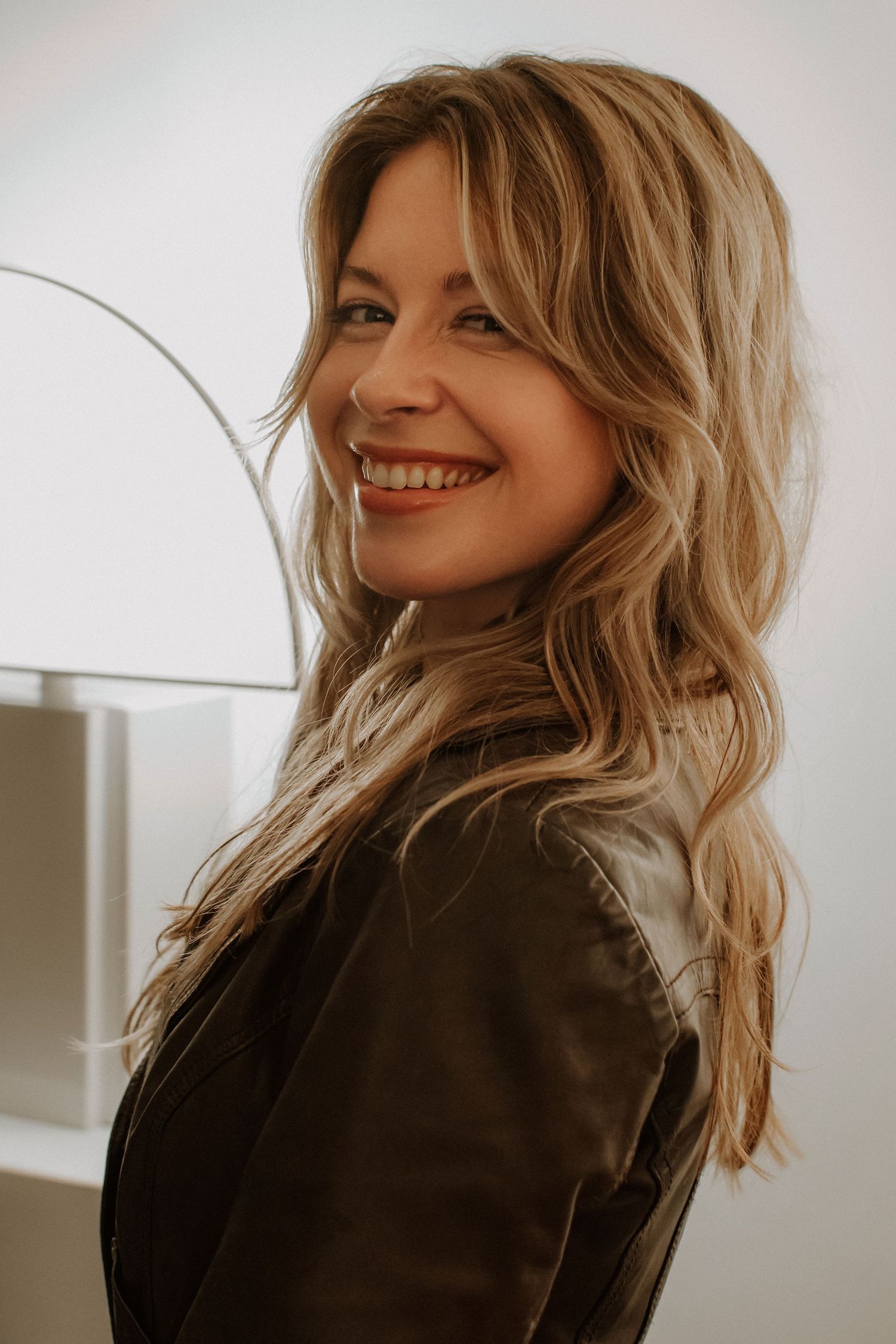 Smiling blonde woman in a dark leather jacket posing in a bright modern interior.