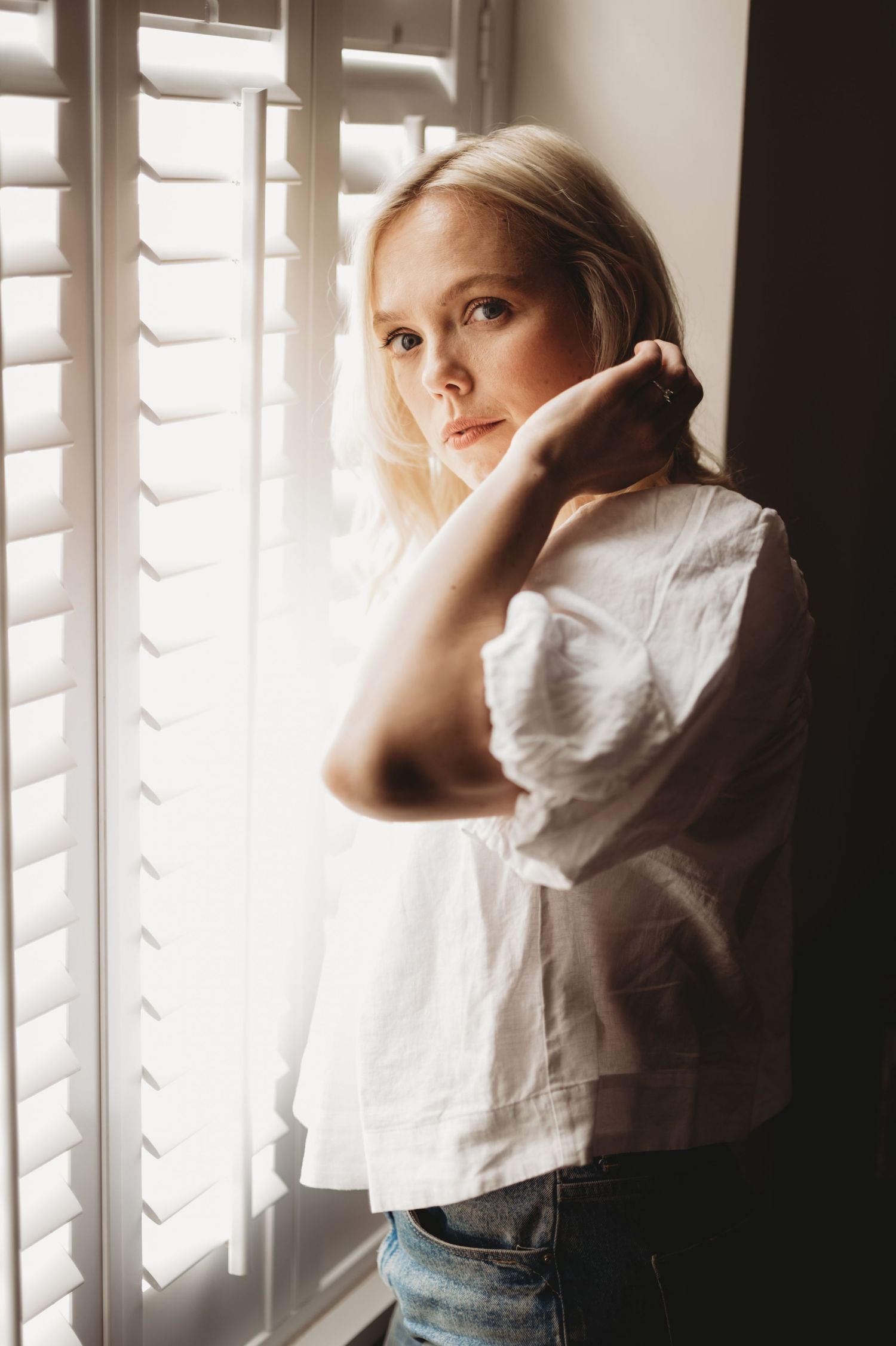 Young blonde woman in white linen top leaning against window shutters in warm natural light.