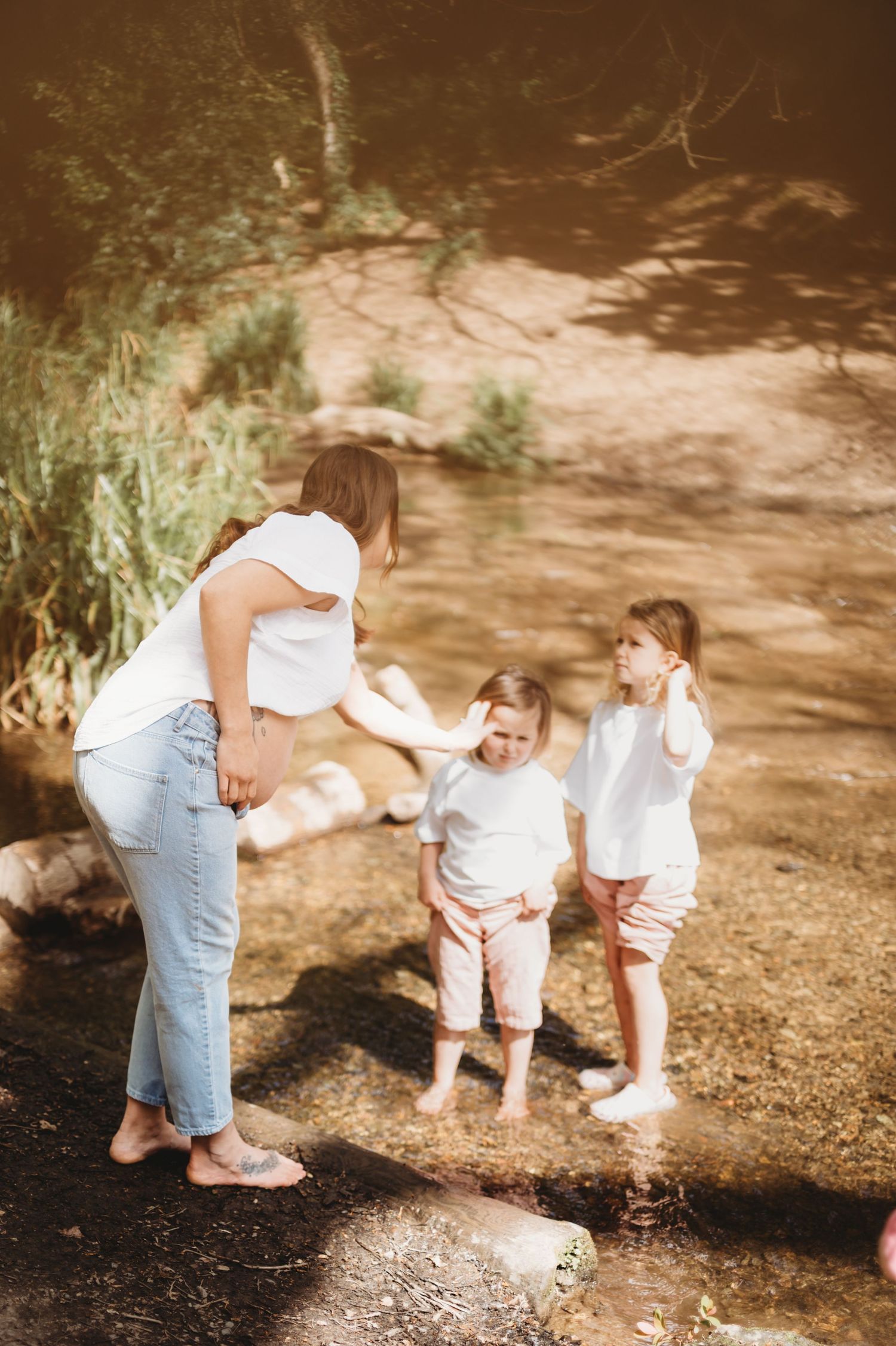 A pregnant mother in white interacts with her two barefoot children playing in a shallow rocky stream.