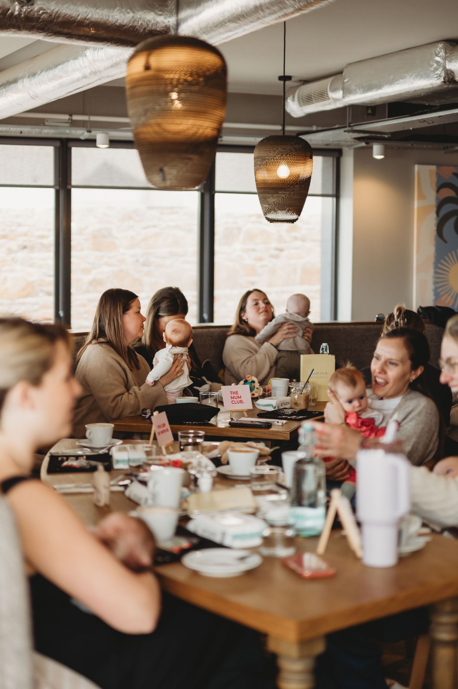 Group of mothers holding babies laughing together at a long wooden table in a cafe.