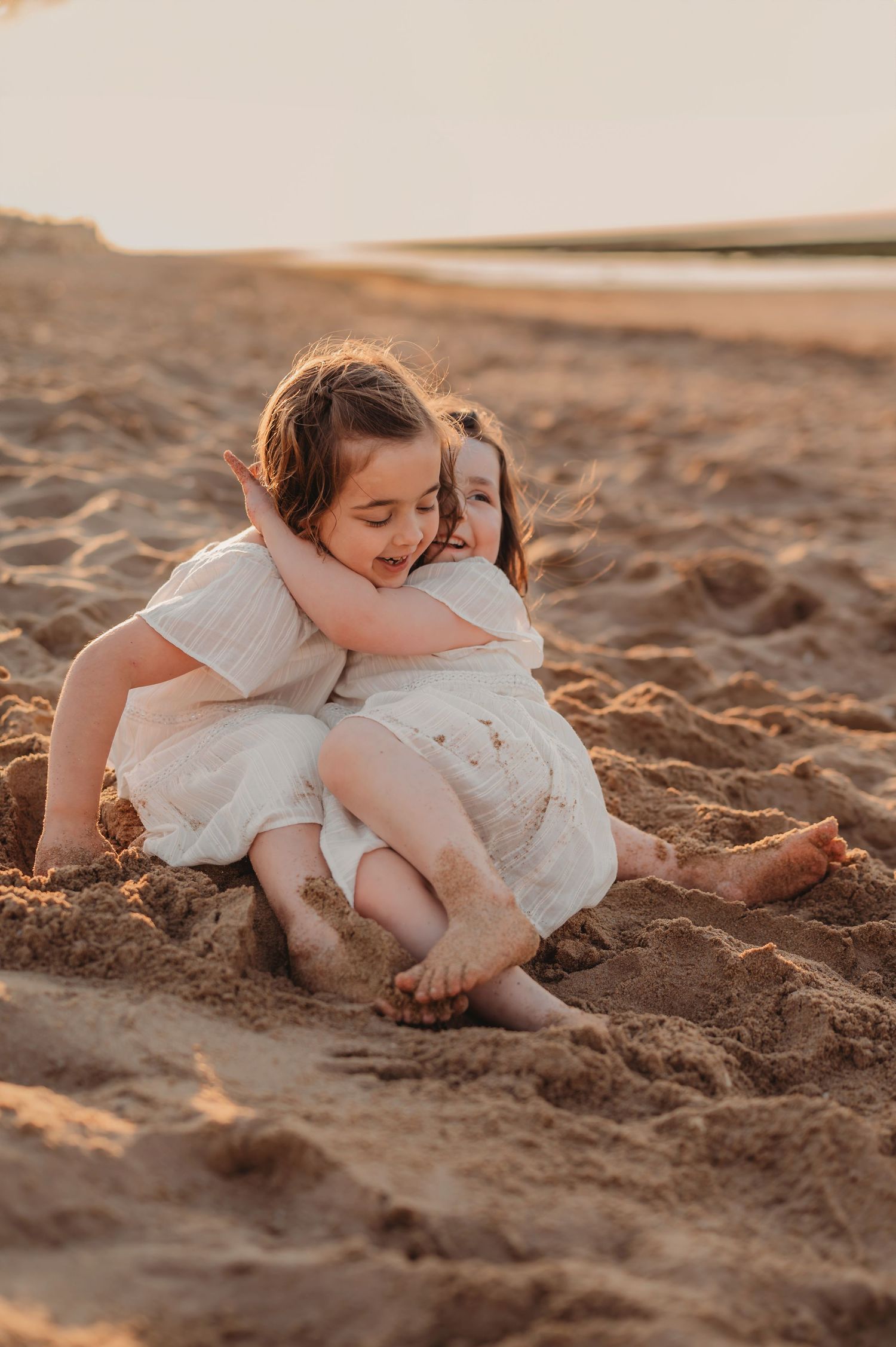 Two young sisters in white dresses hugging and laughing together while sitting in the sand at the beach.