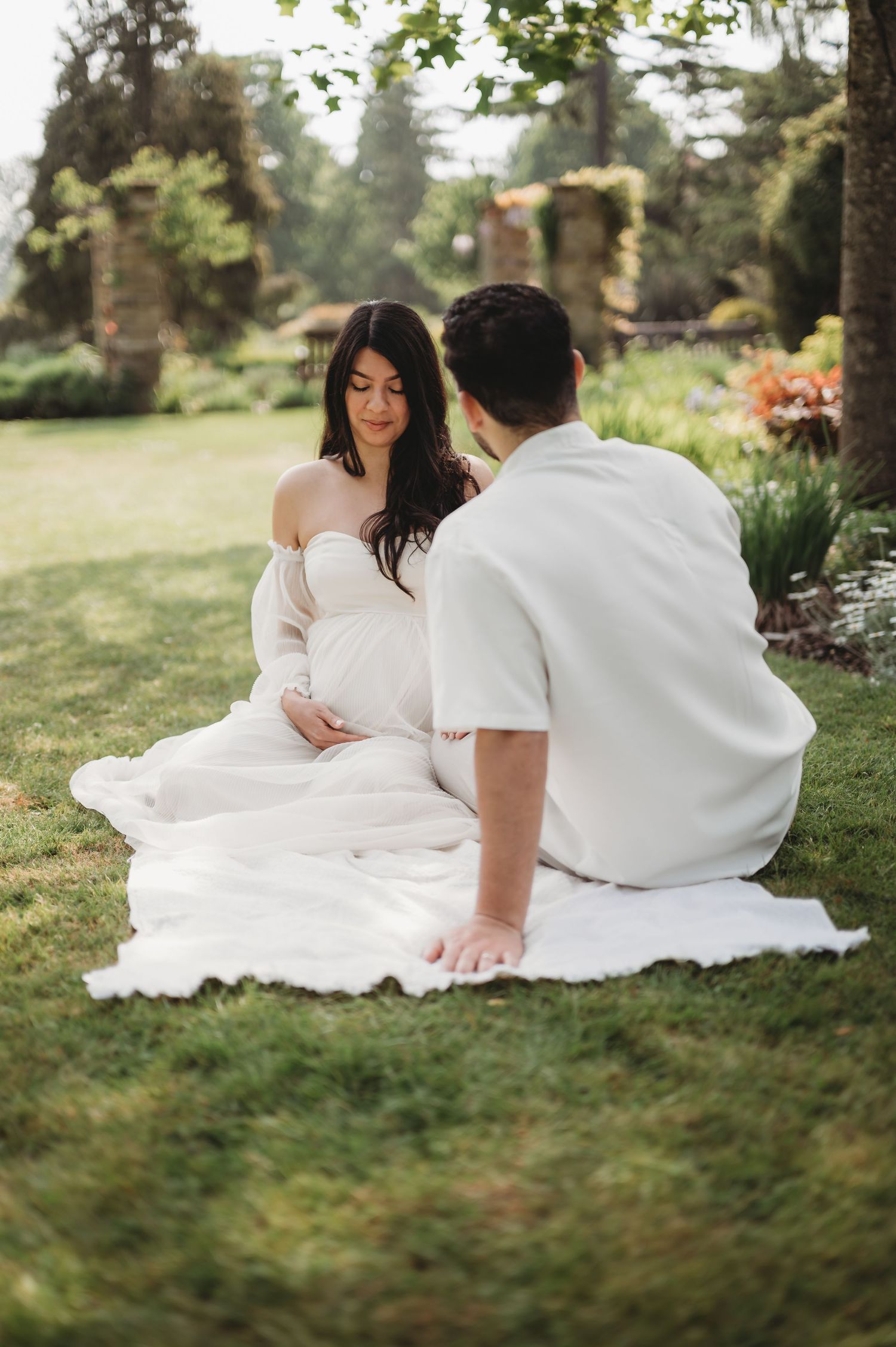 Pregnant woman in flowing white off-shoulder gown sits on grass facing partner in lush garden.