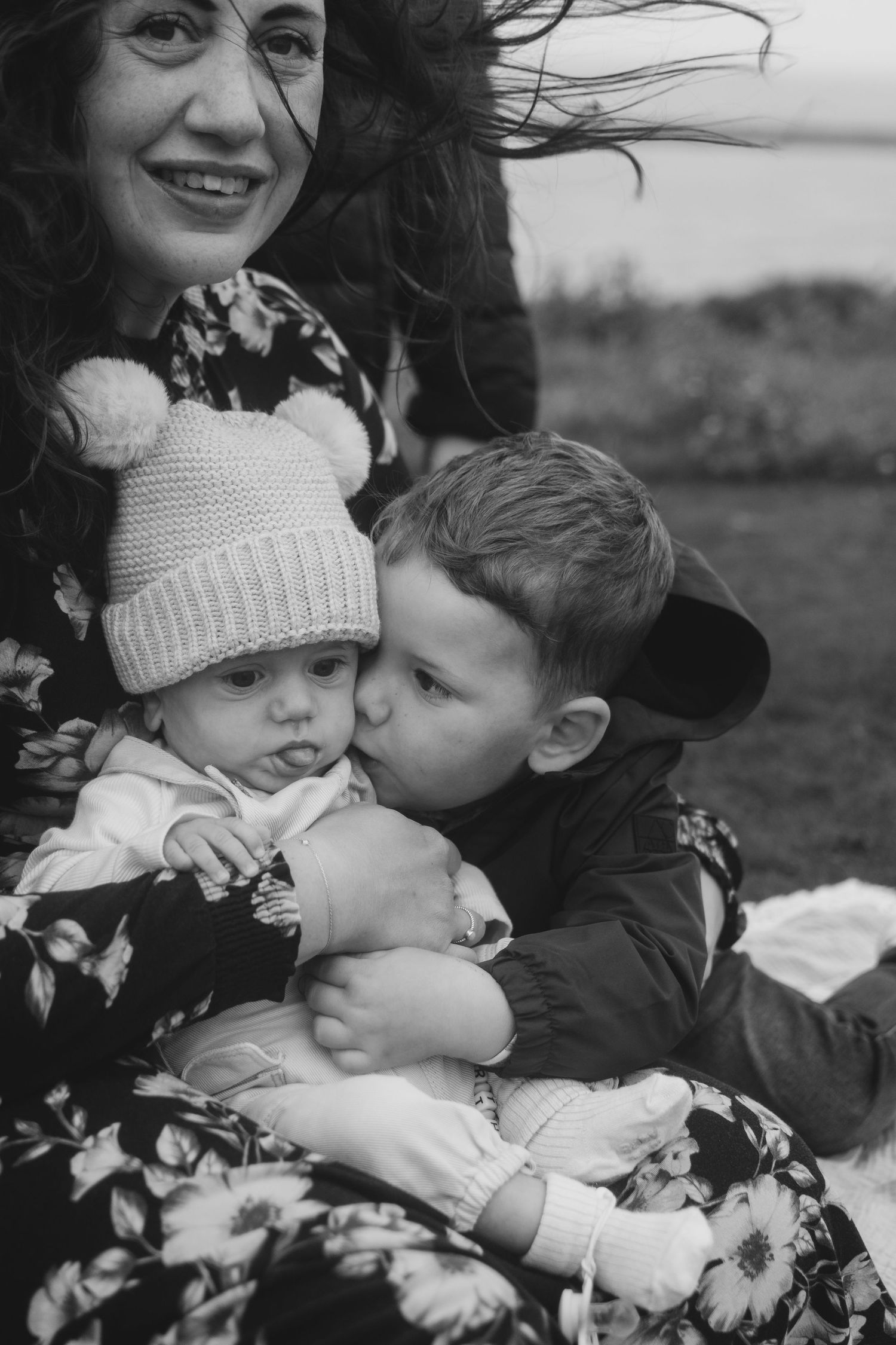 Black and white image of a mother smiling as her son kisses their baby sibling outdoors.