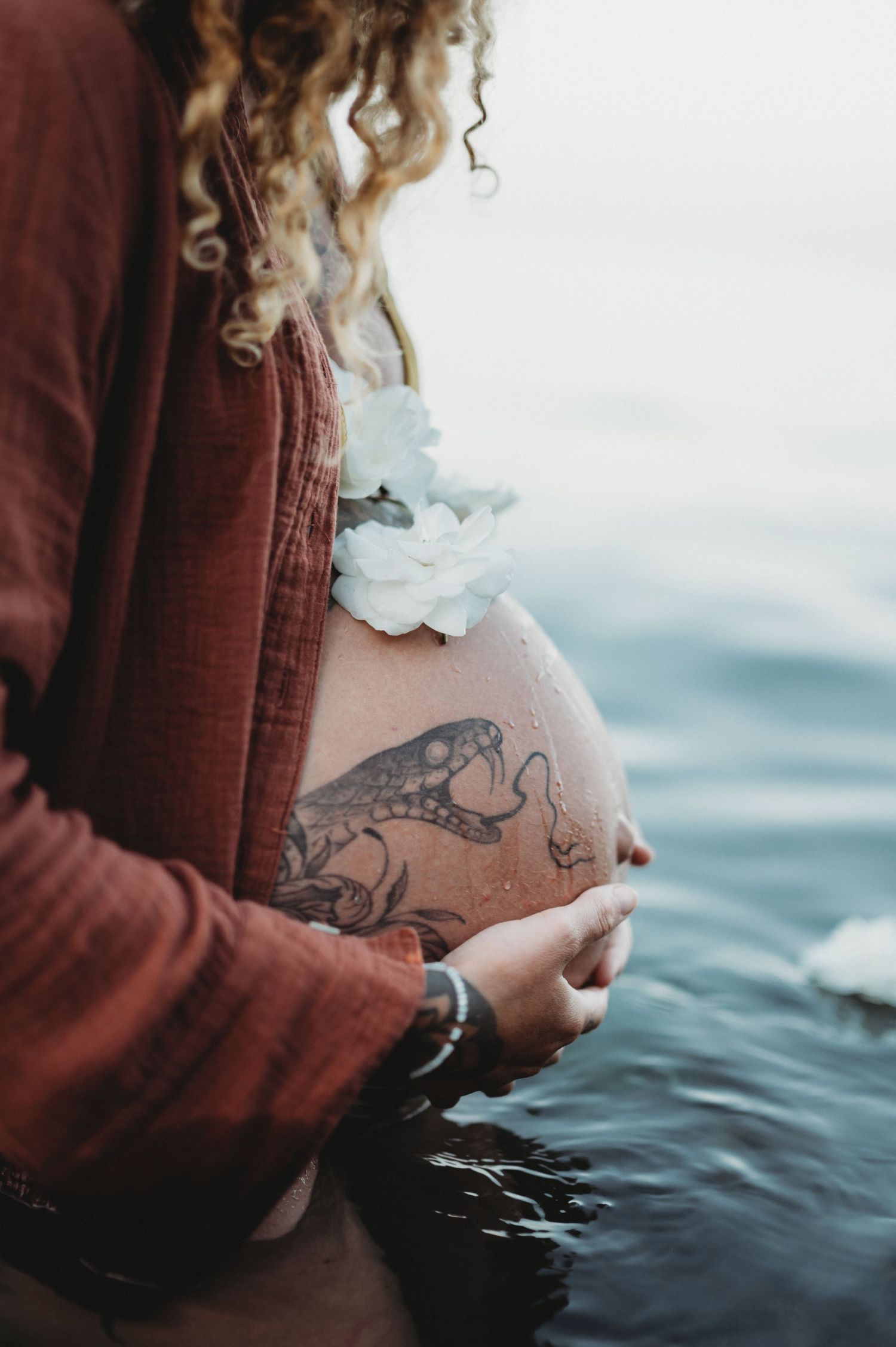Pregnant woman with tattoo cradling bare baby bump near ocean water, wearing red top with white flowers.