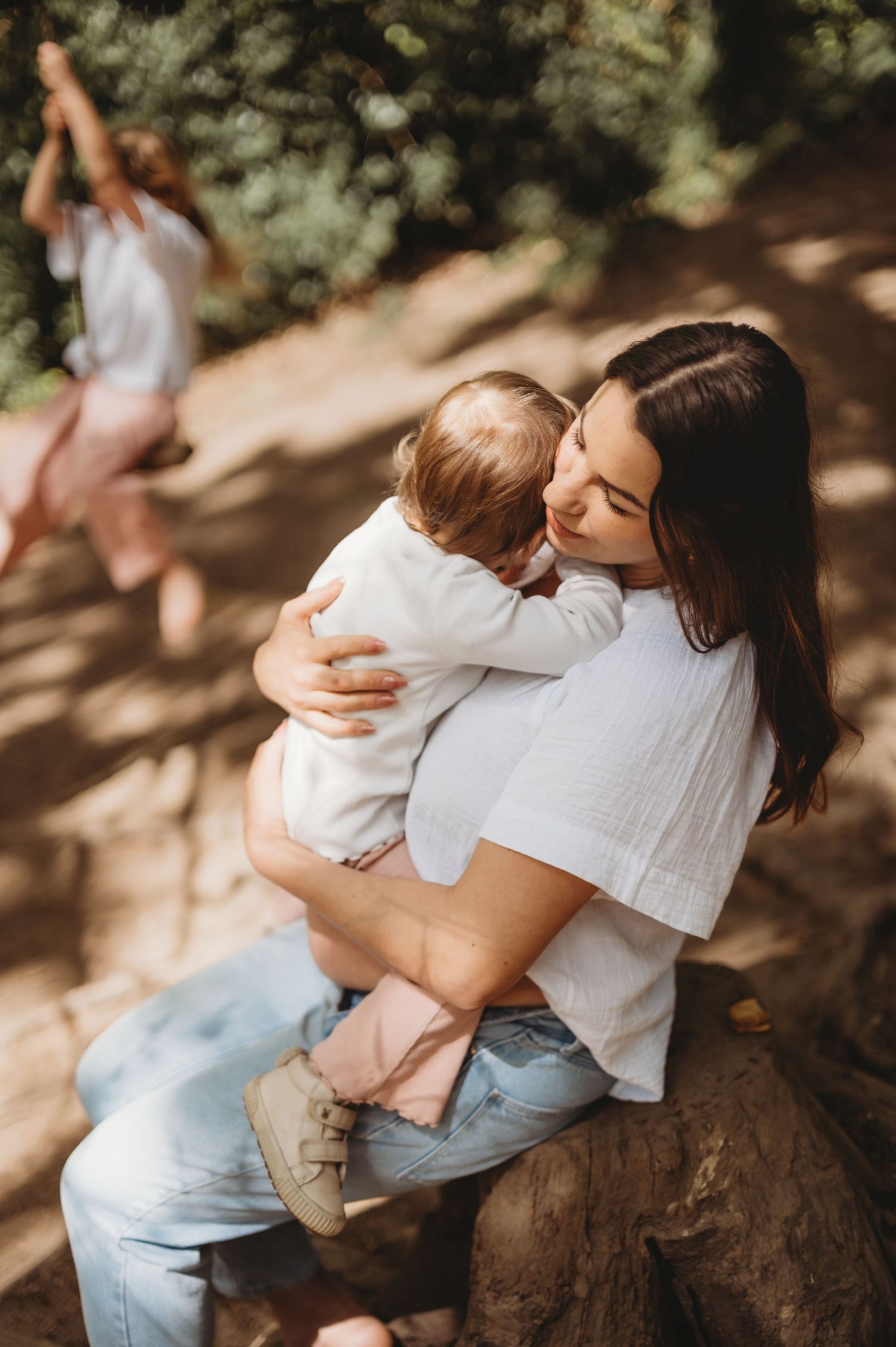 A mother tenderly embraces her toddler while sitting on a rock in a sunlit forest setting.