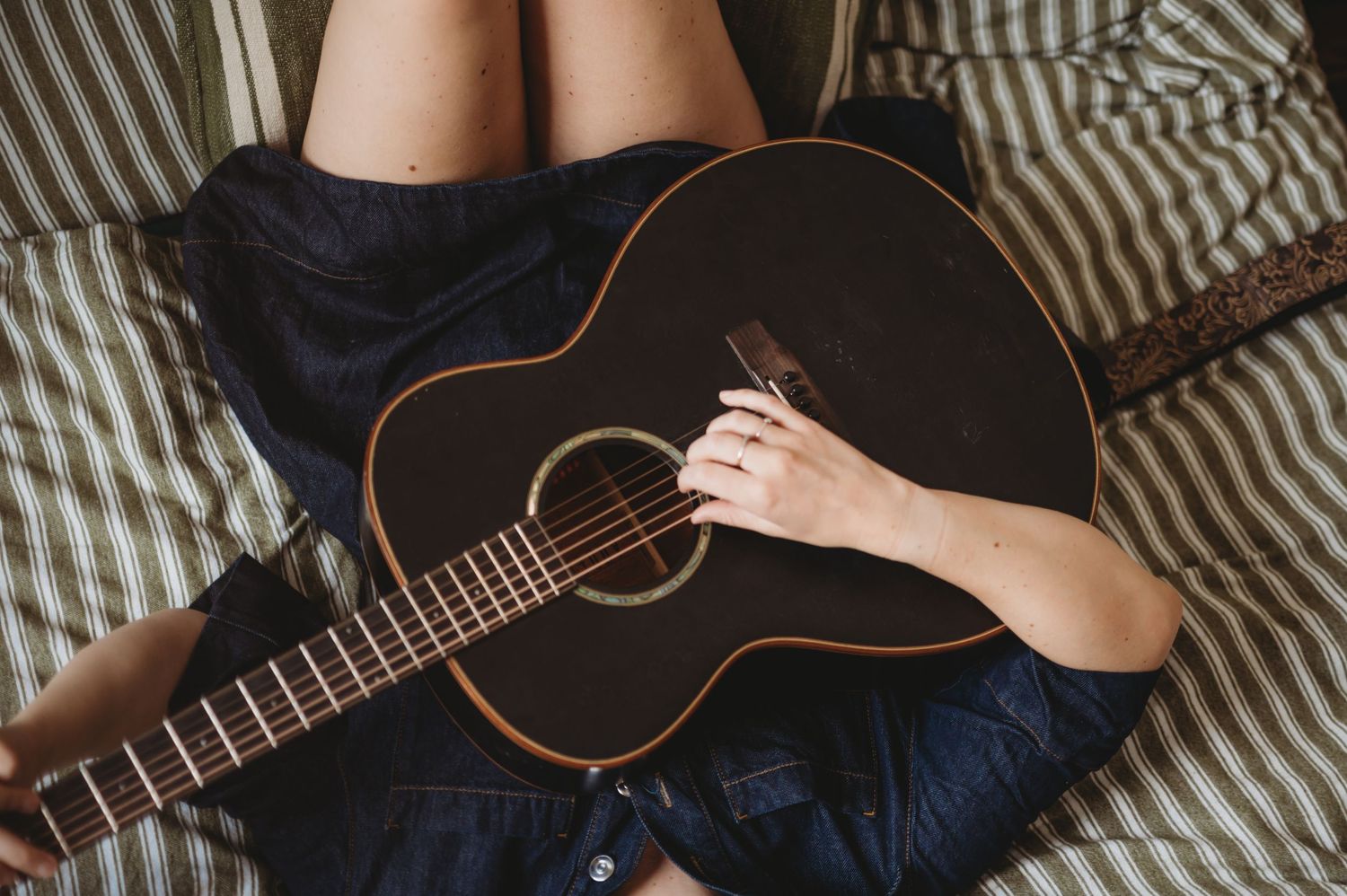 Close-up of a person holding a dark acoustic guitar while sitting on a striped bed.