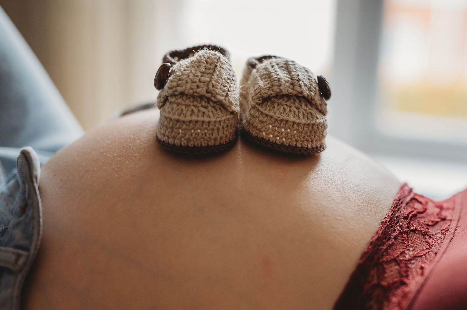 Tiny knitted baby booties resting on pregnant woman's bare baby bump near a window.