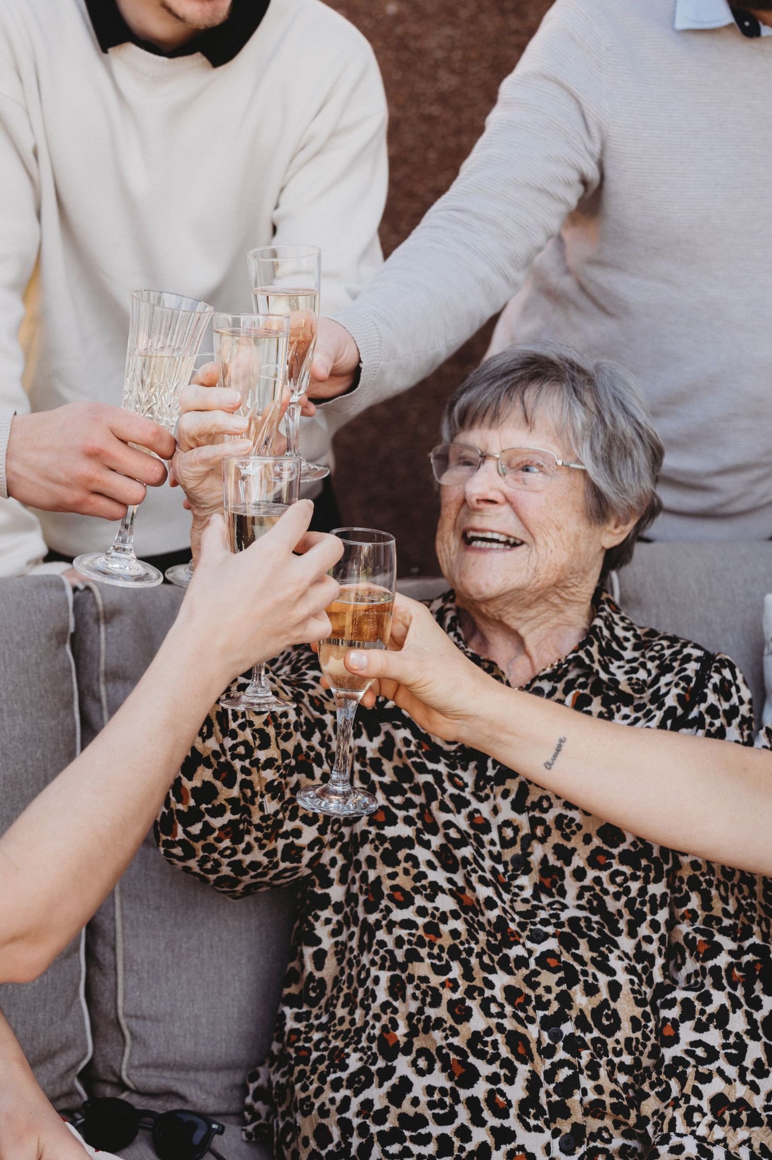An elderly woman laughs joyfully while clinking champagne glasses with family members at a celebration.