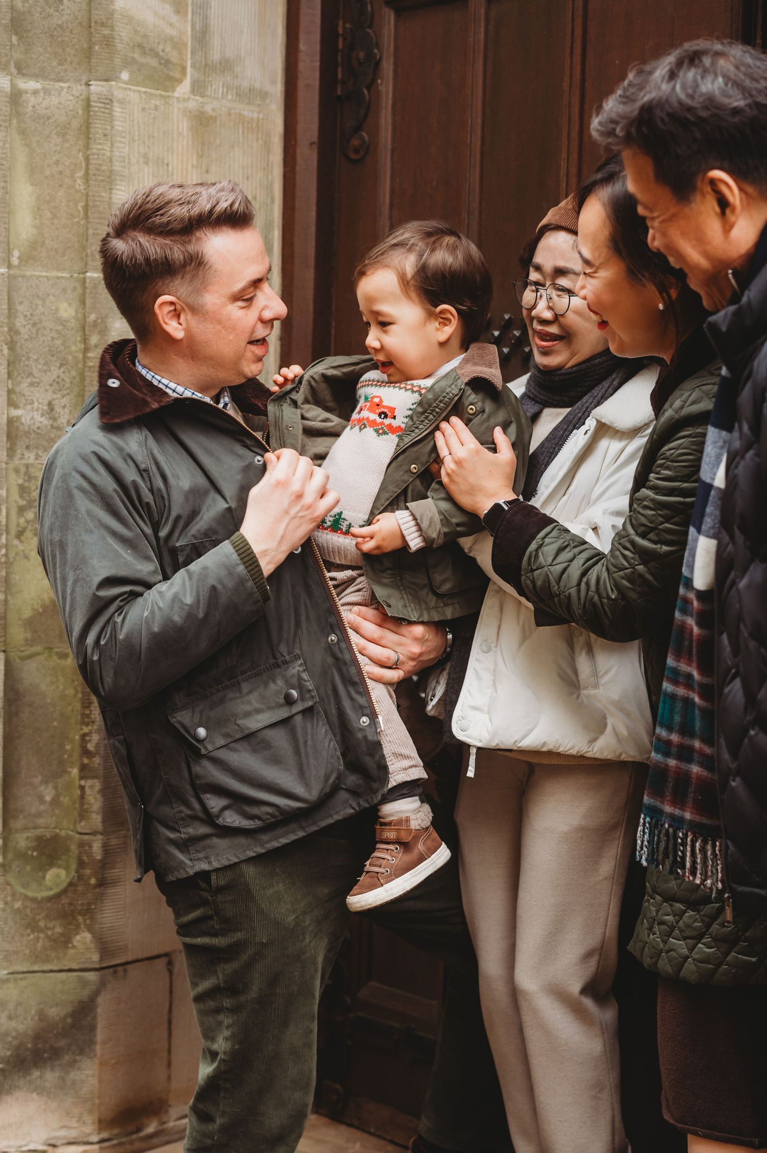 A multigenerational family gathers outdoors smiling and interacting with an adorable baby in winter coats.