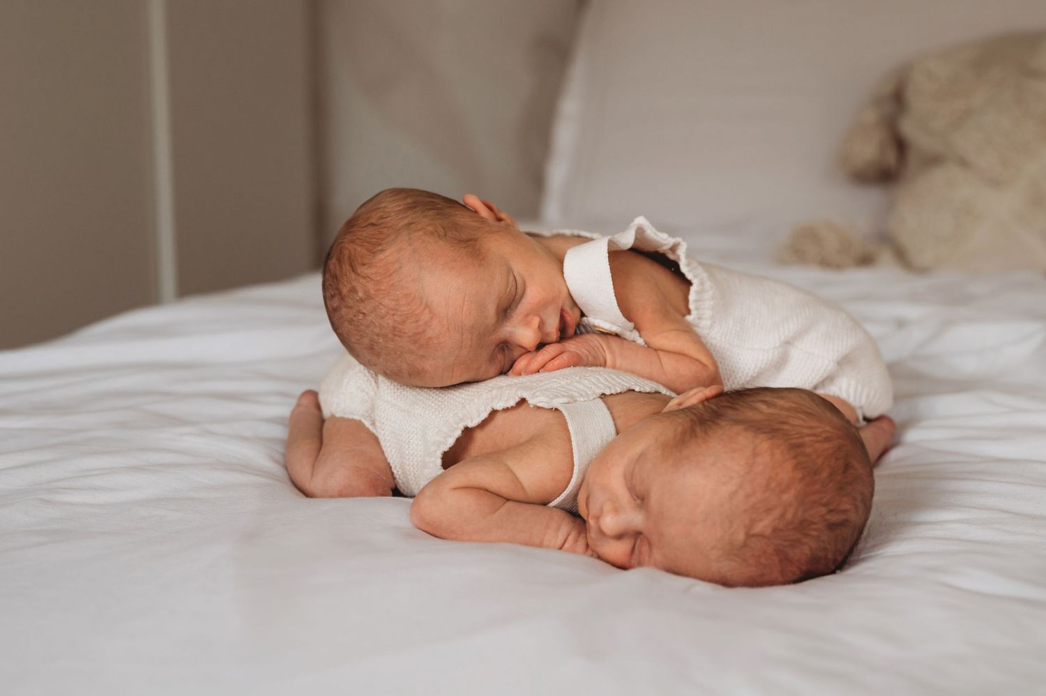 Two sleeping newborn twins in white onesies lying together on soft white bedding.