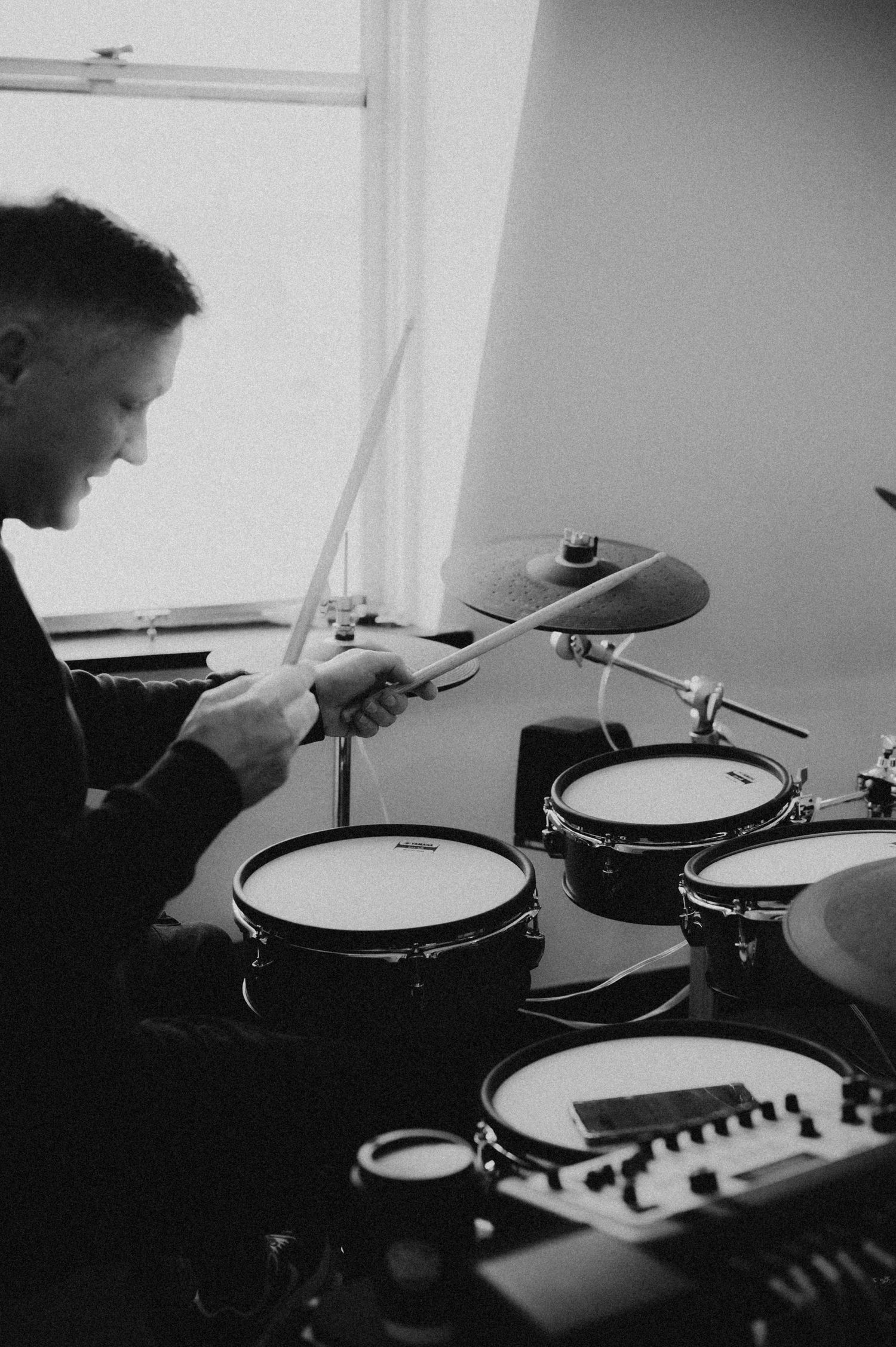A black and white photo of a musician playing an electronic drum kit near a bright studio window.