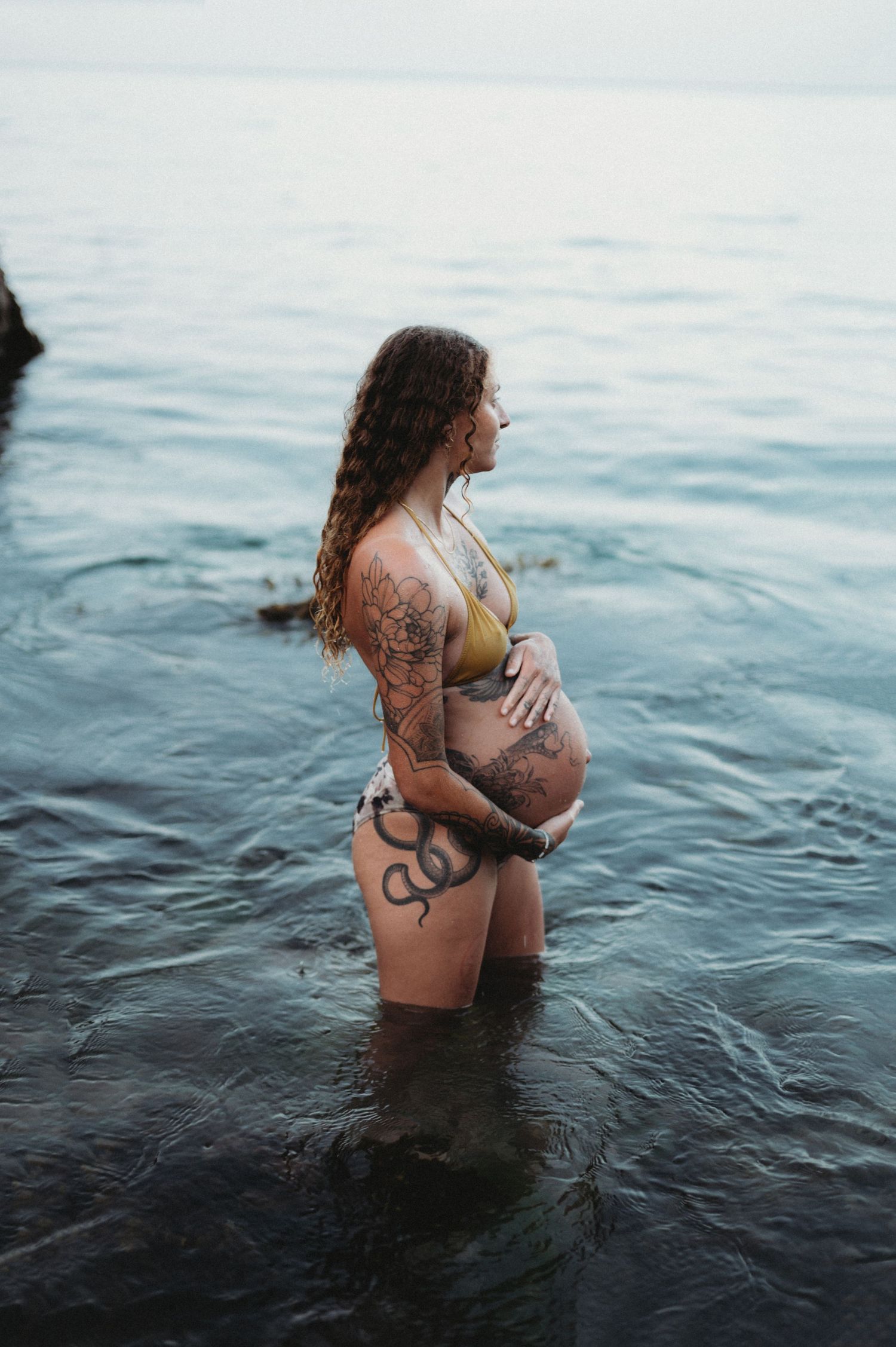Pregnant woman with tattoos in a yellow bikini standing in calm ocean water, cradling her baby bump.