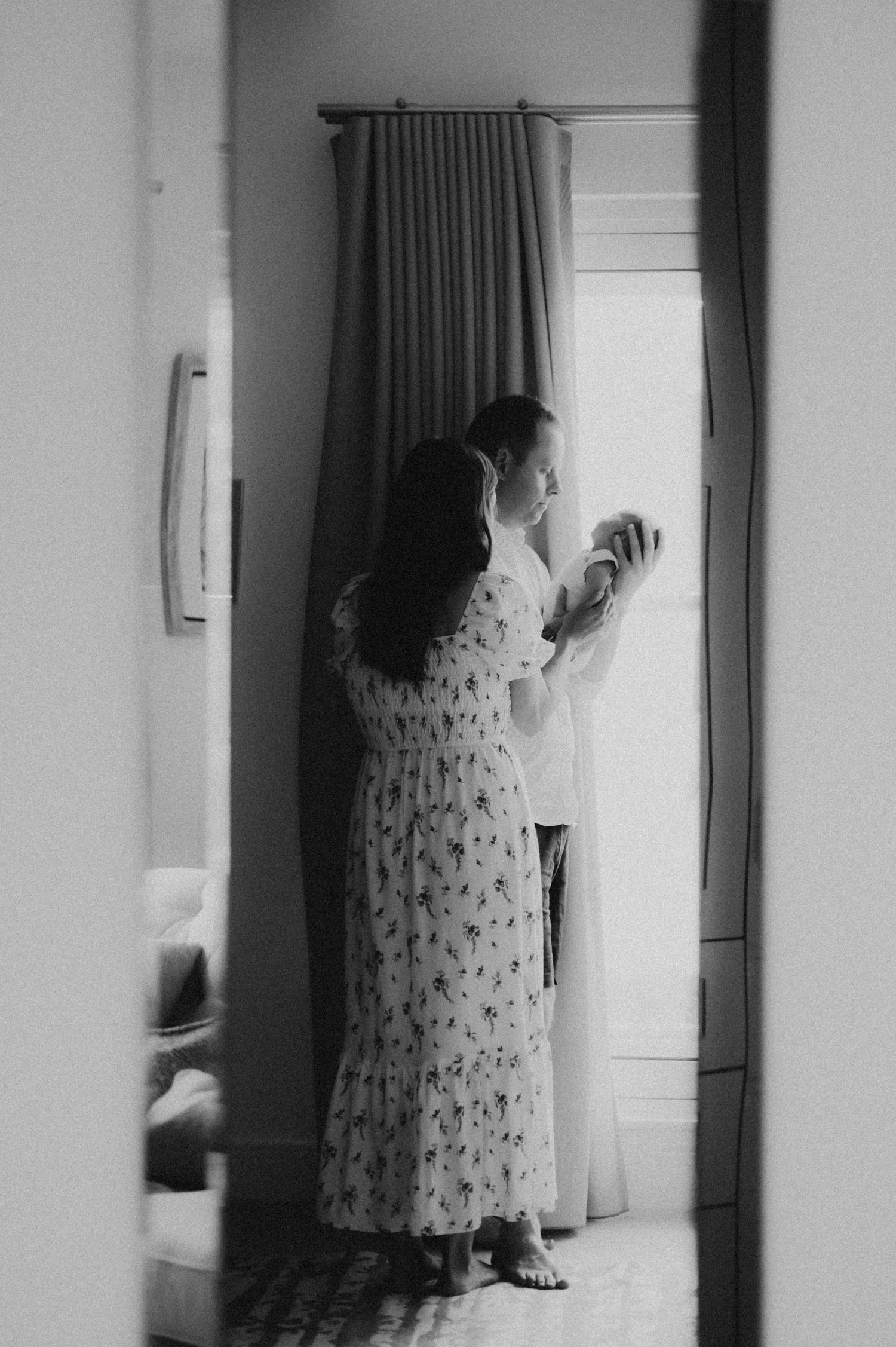 Black and white photo of a couple standing by a curtained window, woman in floral dress, man holding a newborn baby.