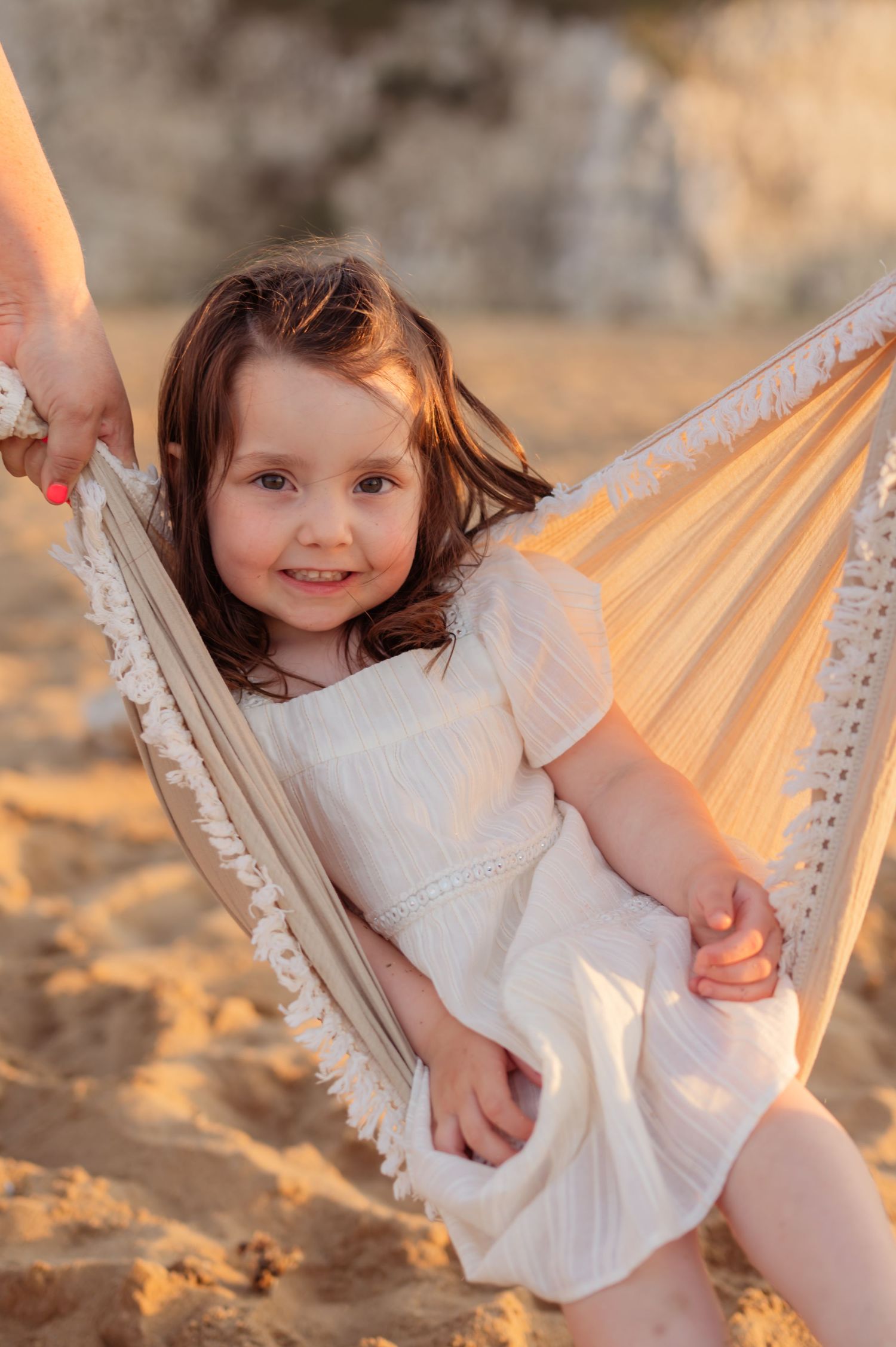 Smiling little girl in white dress sitting in a hammock on a sandy beach at golden hour.