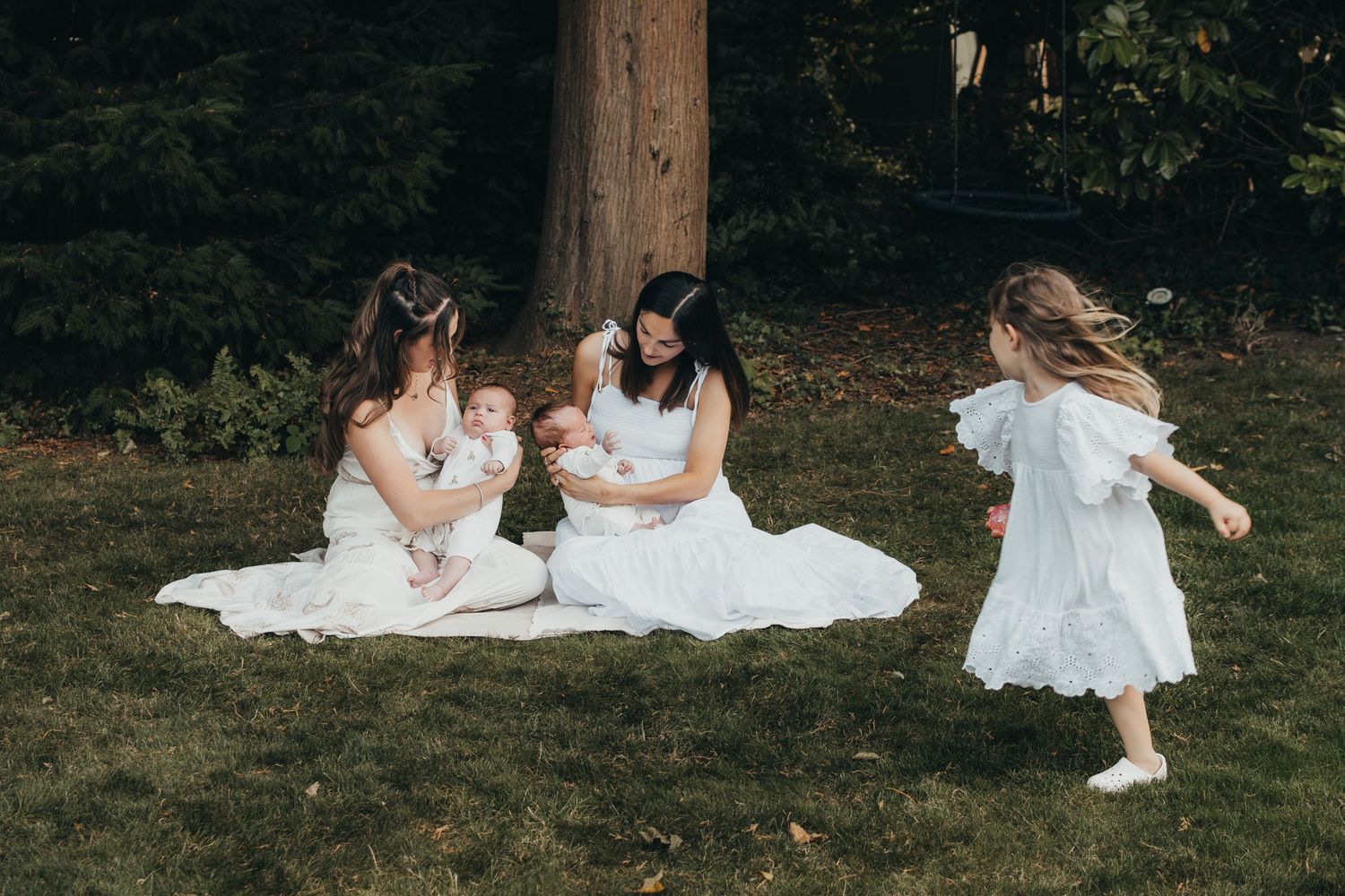 Two women in white dresses sitting on a blanket holding babies while a young girl twirls nearby.