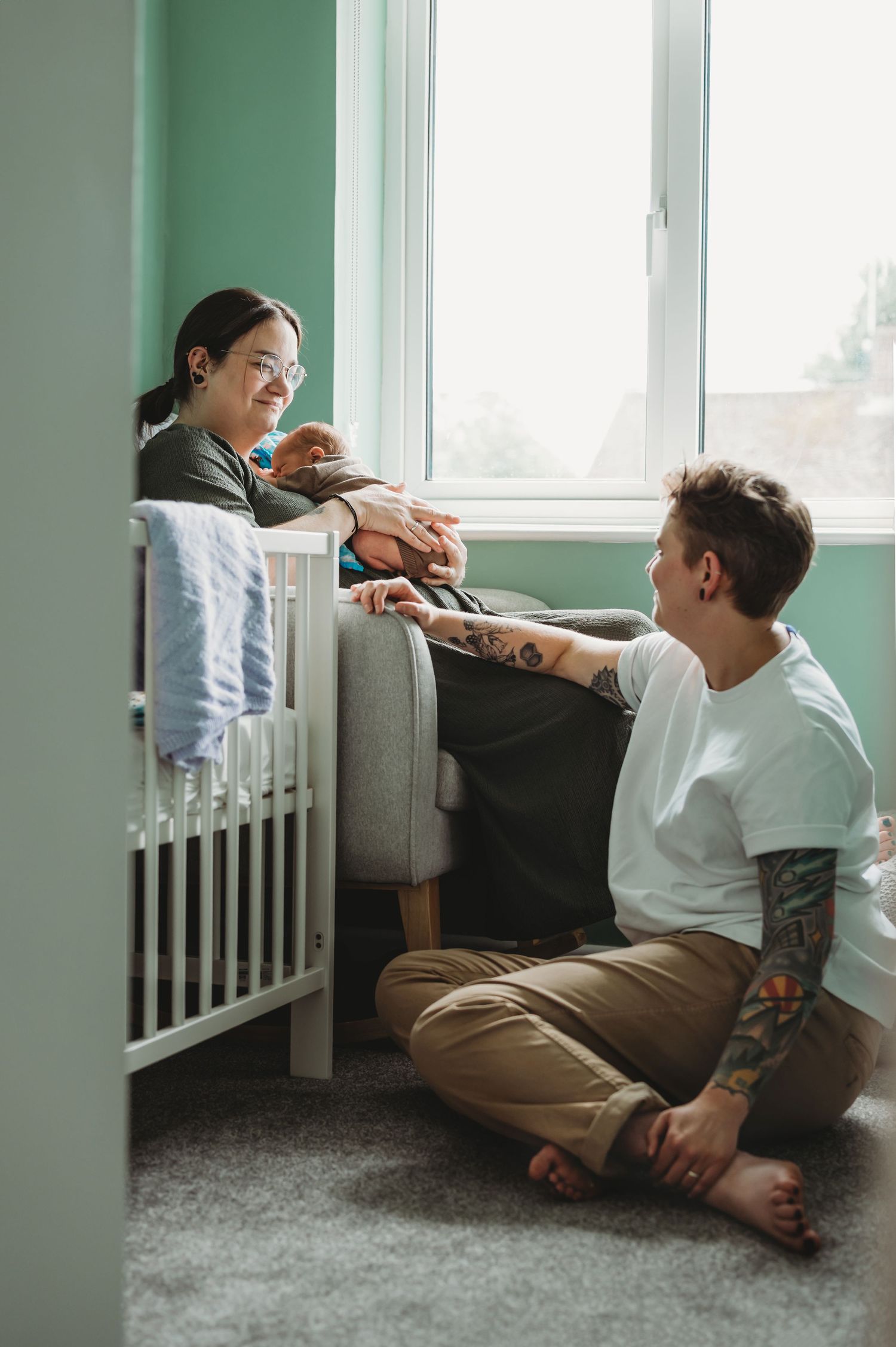 Two parents share a tender moment with their newborn near a white crib by a window.