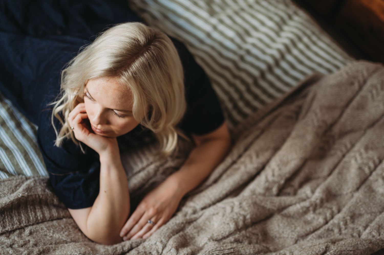 Blonde woman in navy top lying on a bed with striped pillows, looking thoughtful.