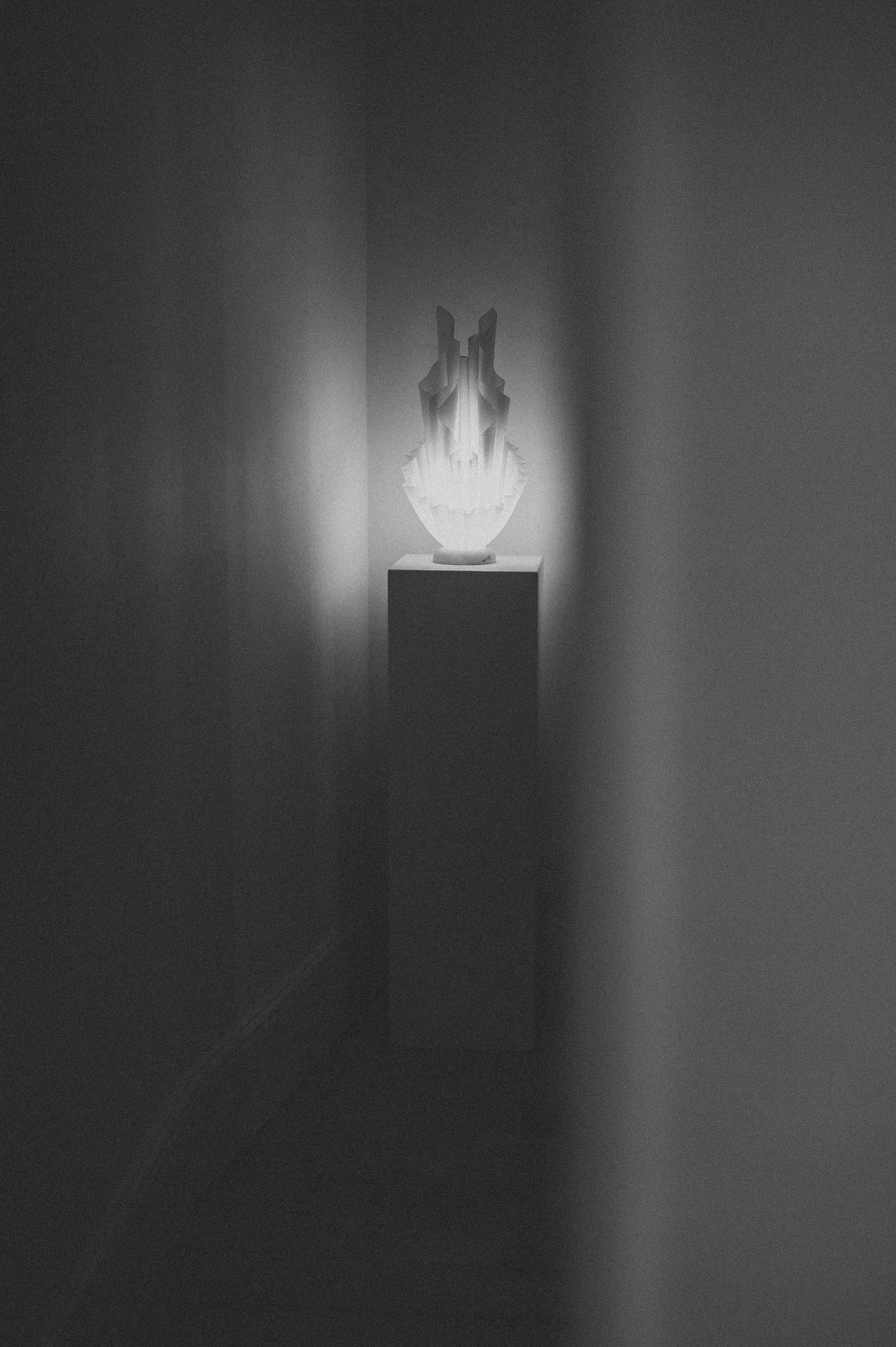 A glowing white sculptural vase displayed on a dark pedestal in a dimly lit gallery corridor.
