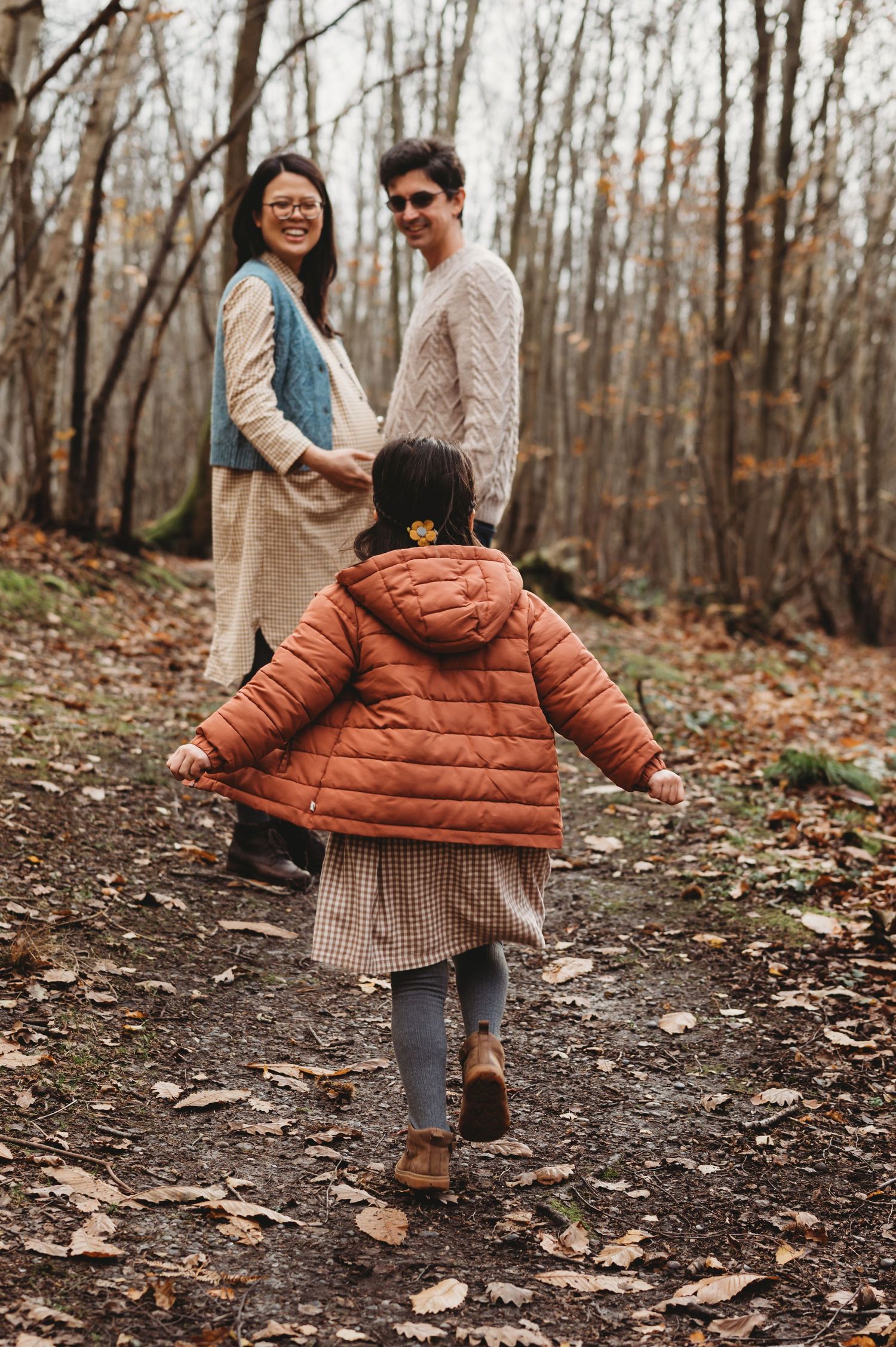 A child in an orange puffer jacket runs toward her parents on a leaf-covered autumn forest path.