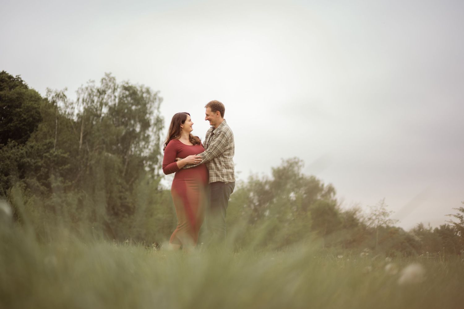 Couple standing together in lush green field during maternity photoshoot.