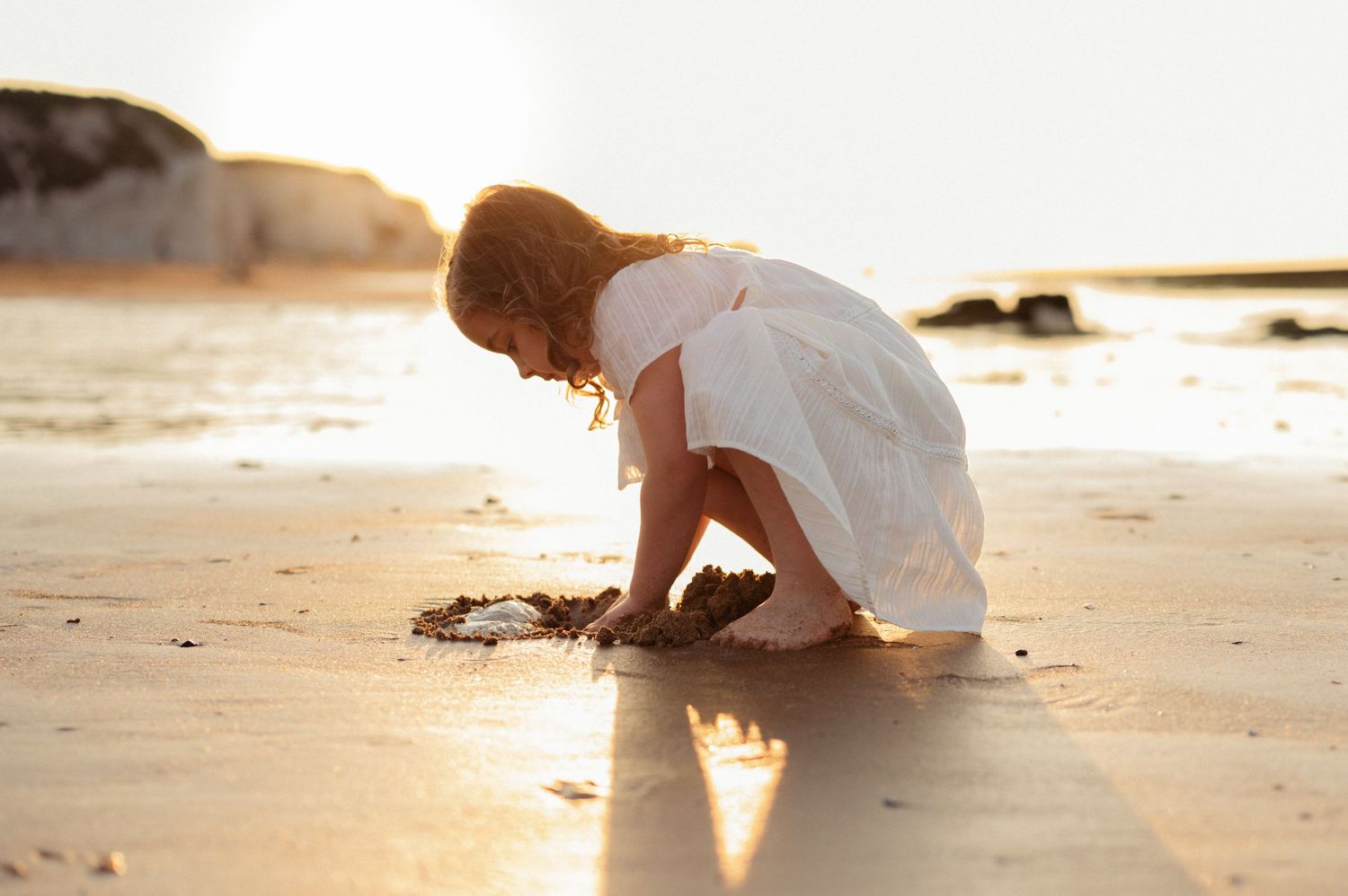 Young girl in white dress crouching on wet sand drawing shapes at the beach during sunset.