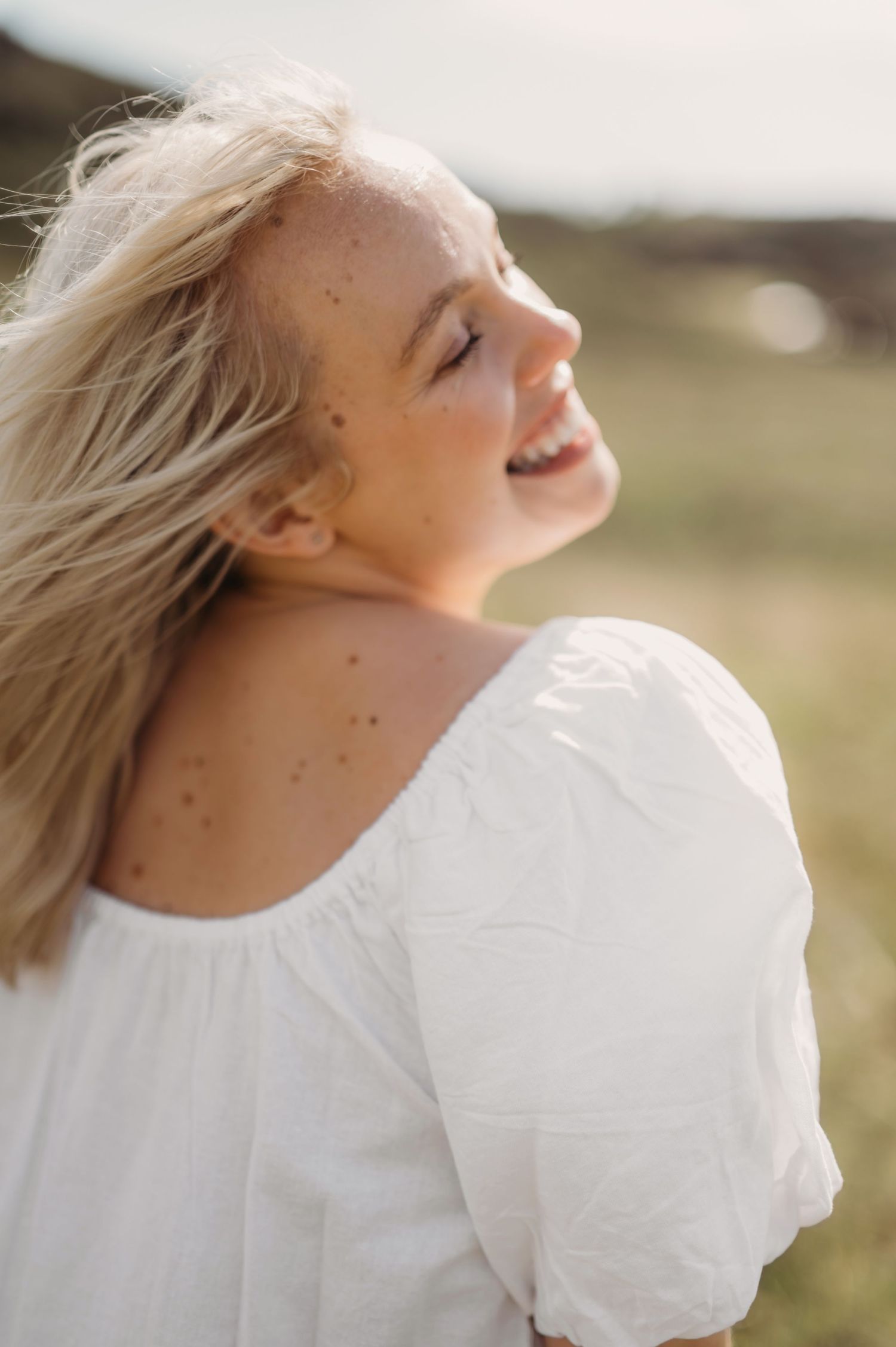 Smiling blonde woman with freckles in white off-shoulder dress enjoying sunshine outdoors.
