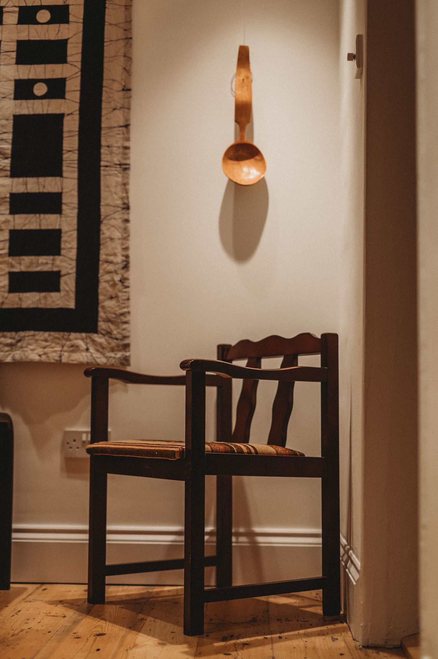 Dark wooden armchair beneath a sculptural wooden wall sconce beside a woven textile wall hanging.